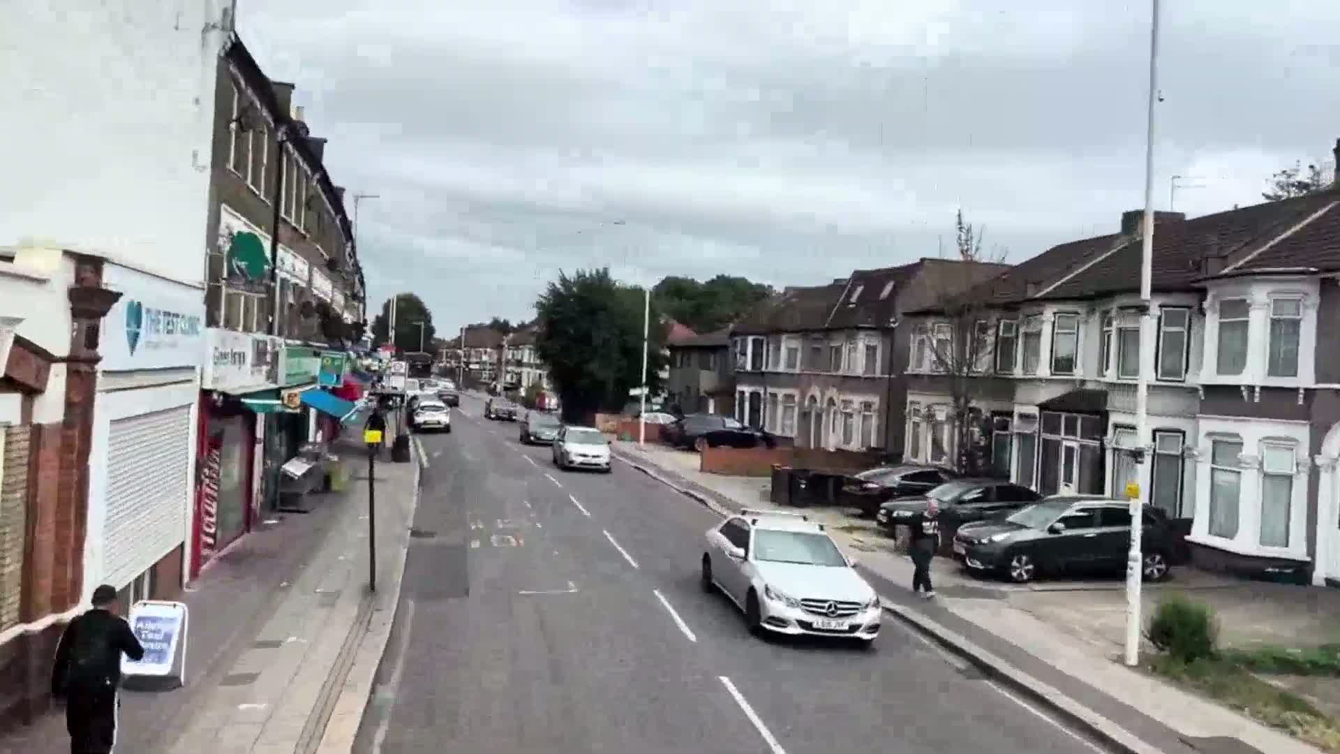 A residential street lined with shops and houses on a cloudy day, with several cars driving along the road and a person walking on the sidewalk.