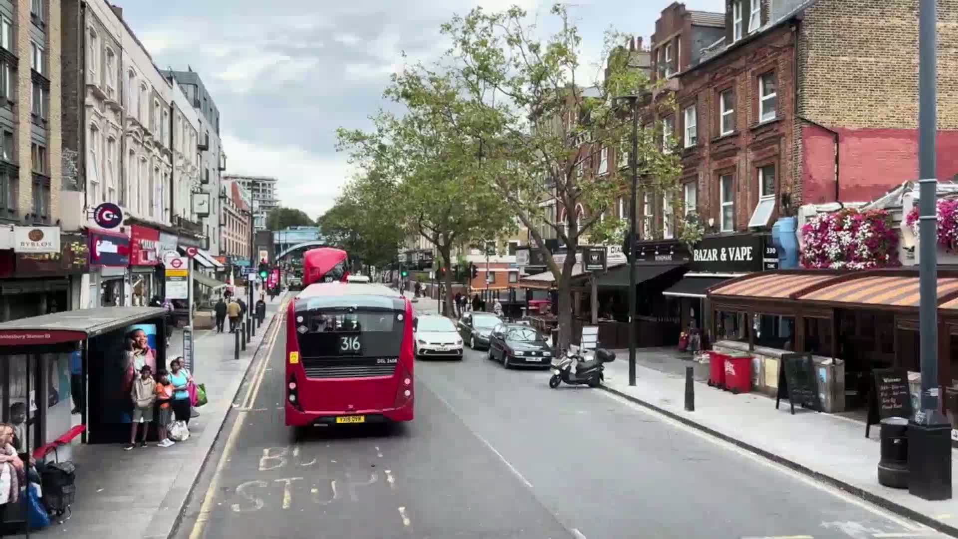 A red double-decker bus drives down a city street on an overcast day, passing pedestrians at a bus stop and cars parked along the side of the road lined with shops and restaurants.