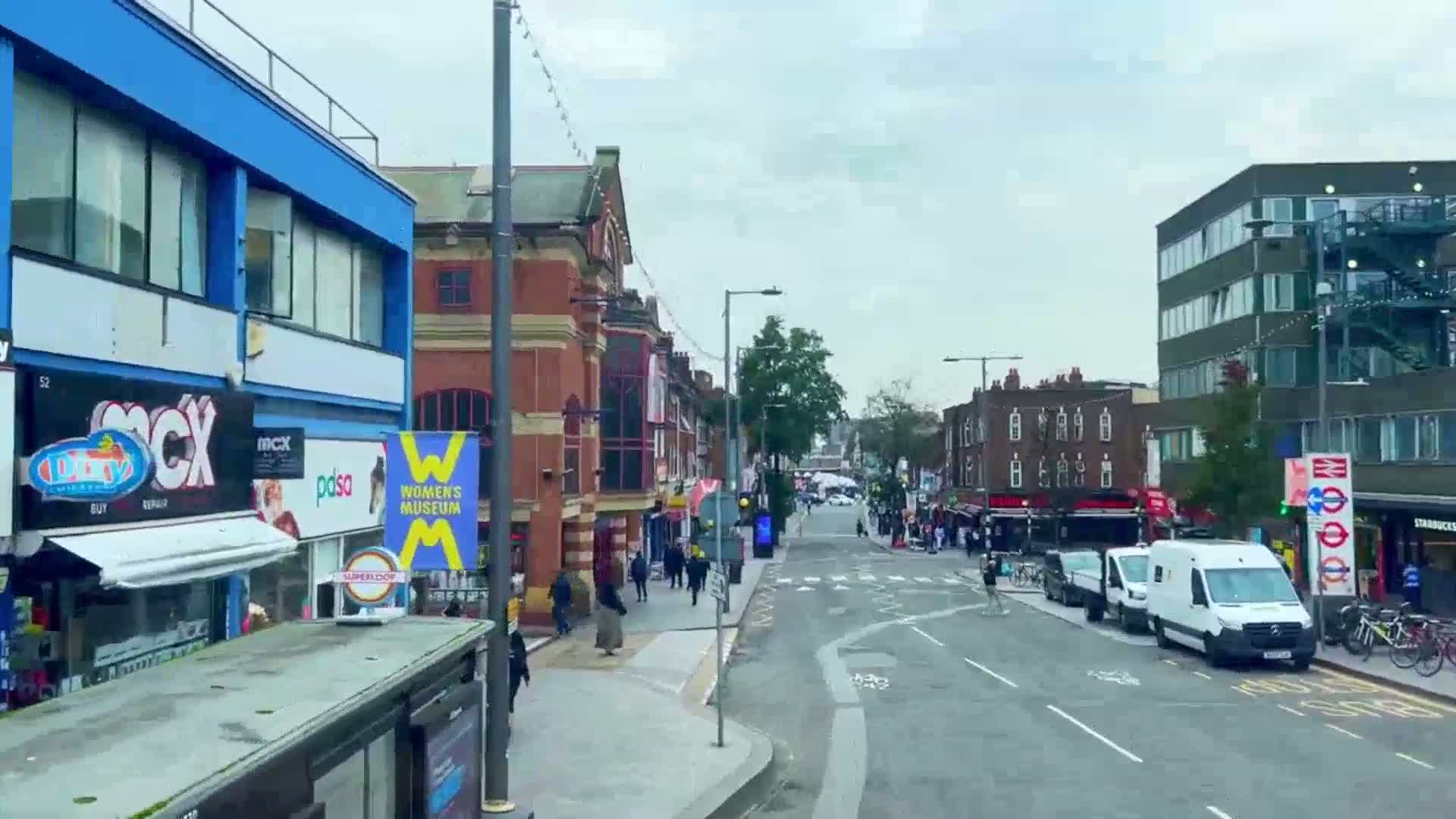 A city street scene on an overcast day shows multi-story buildings with shops and pedestrians, with vehicles parked and moving along the roadway.