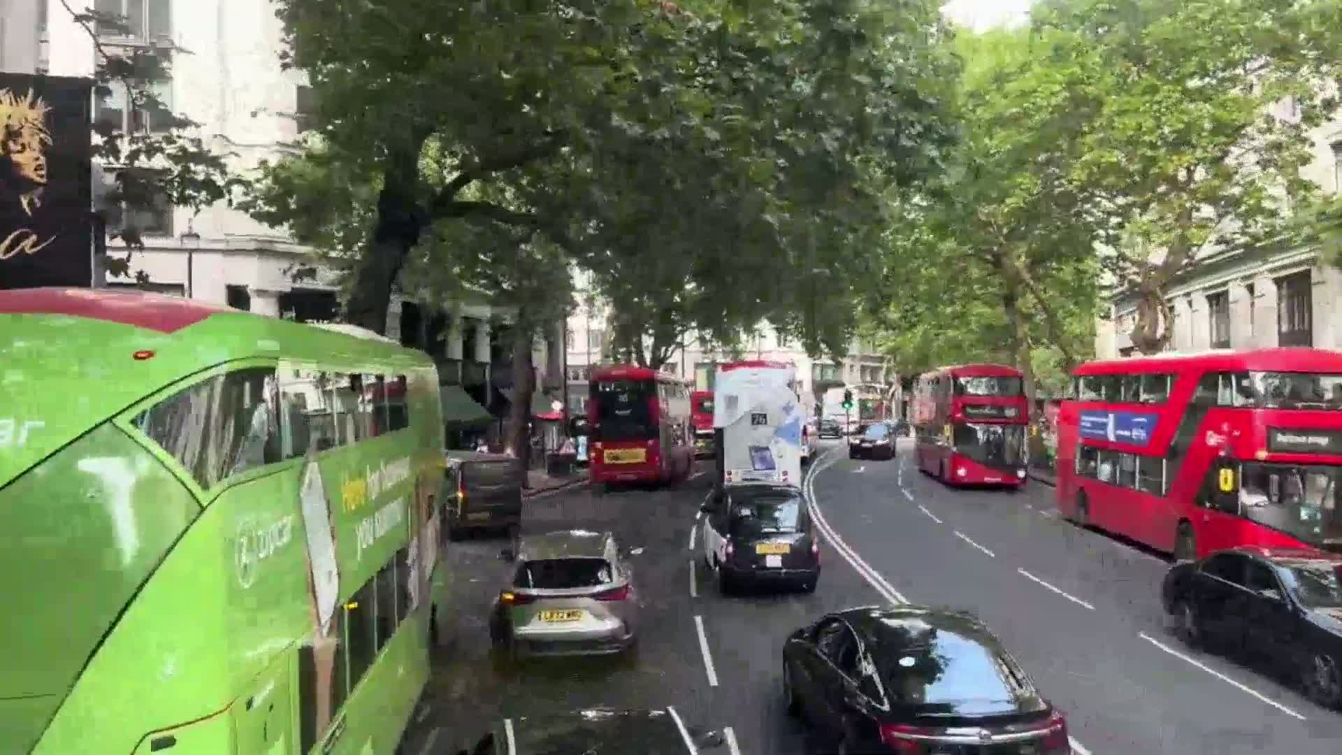A green double-decker bus drives past other buses and cars on a city street under a partly sunny sky.