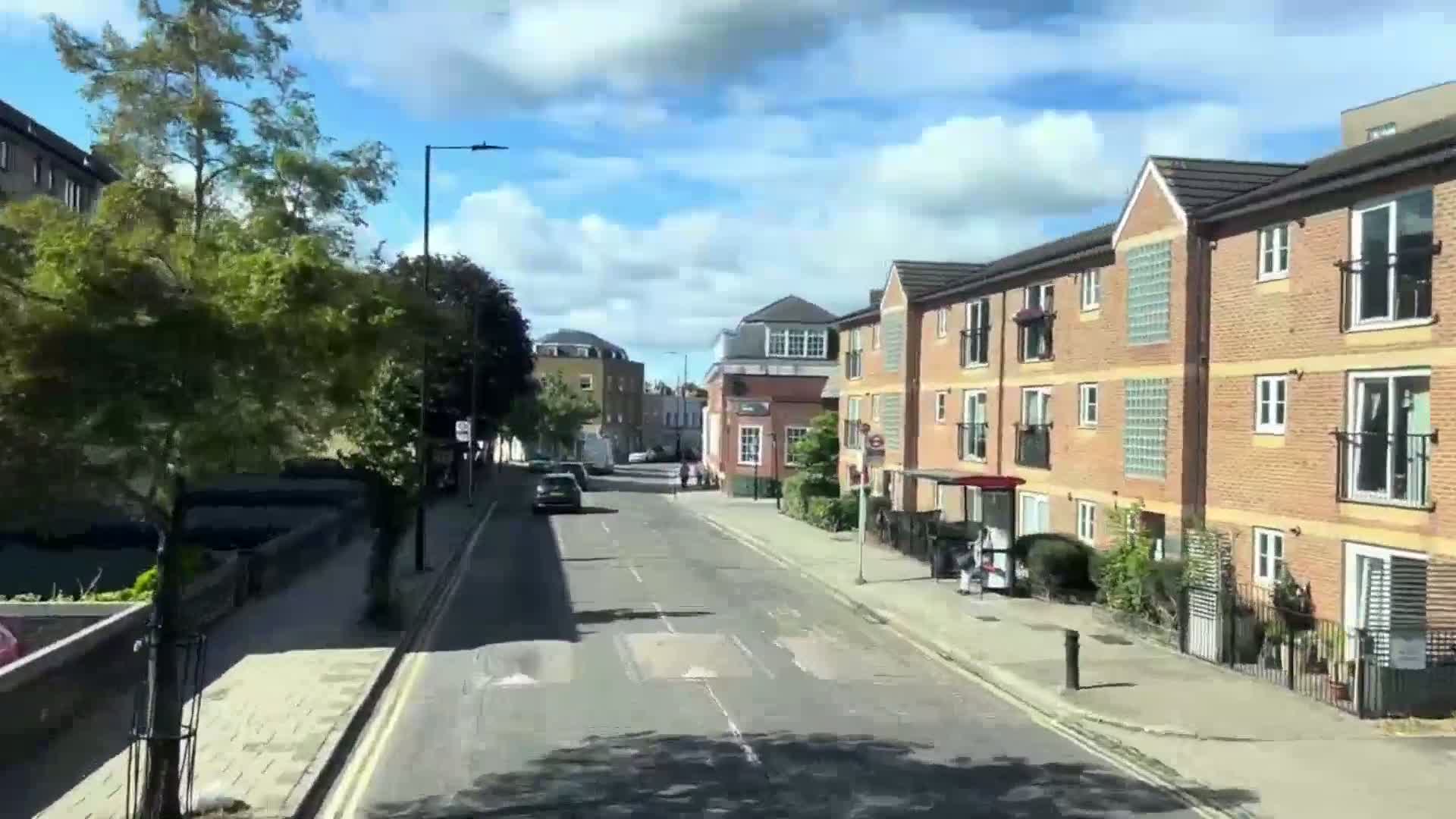 A sunny day shows a residential street with two- and three-story brick buildings, trees lining the sidewalks, and a few cars parked and driving.