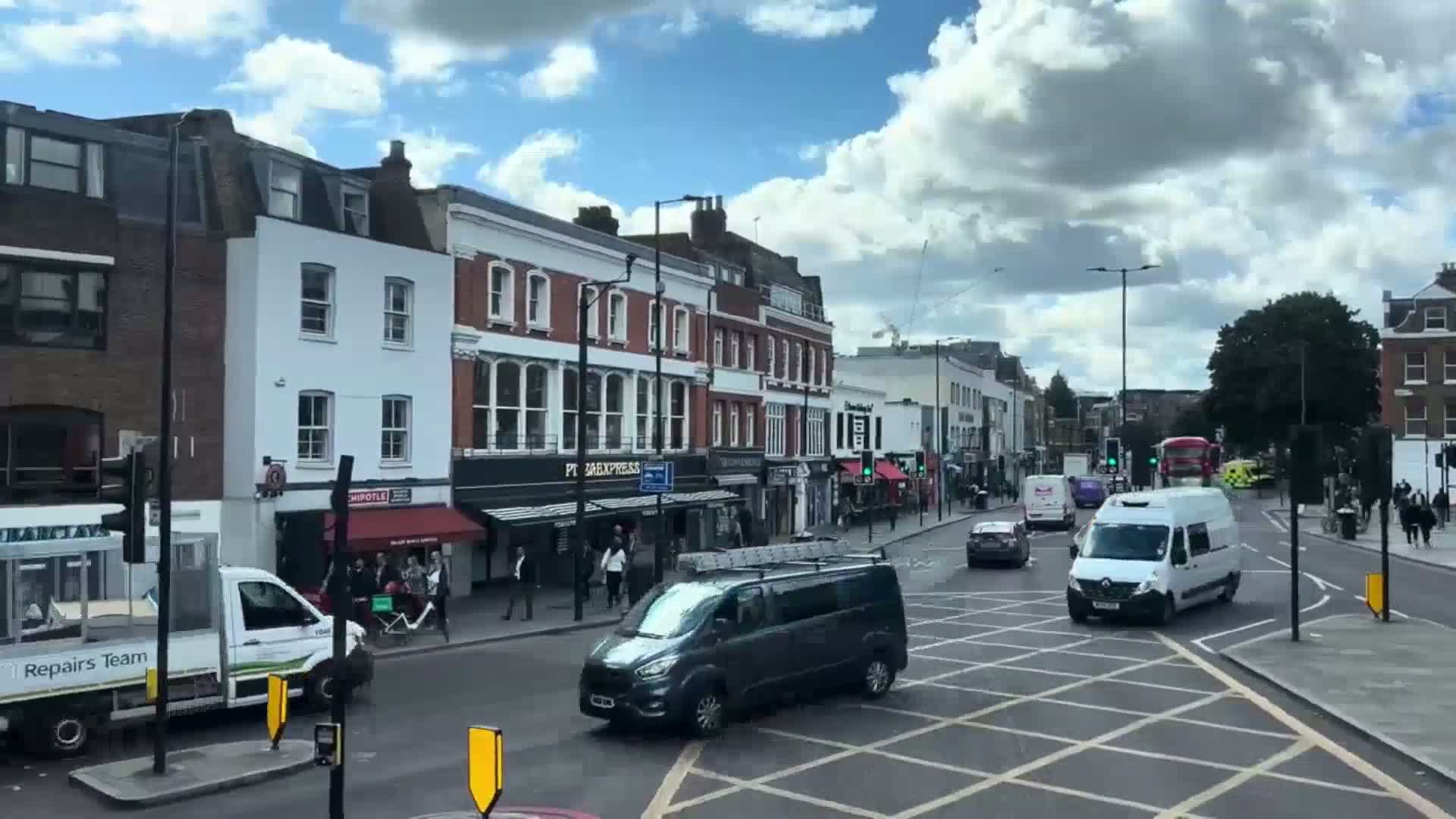 A partly cloudy day reveals a busy street scene in a city with multi-story brick buildings, shops, pedestrians, and several vans and cars at an intersection.