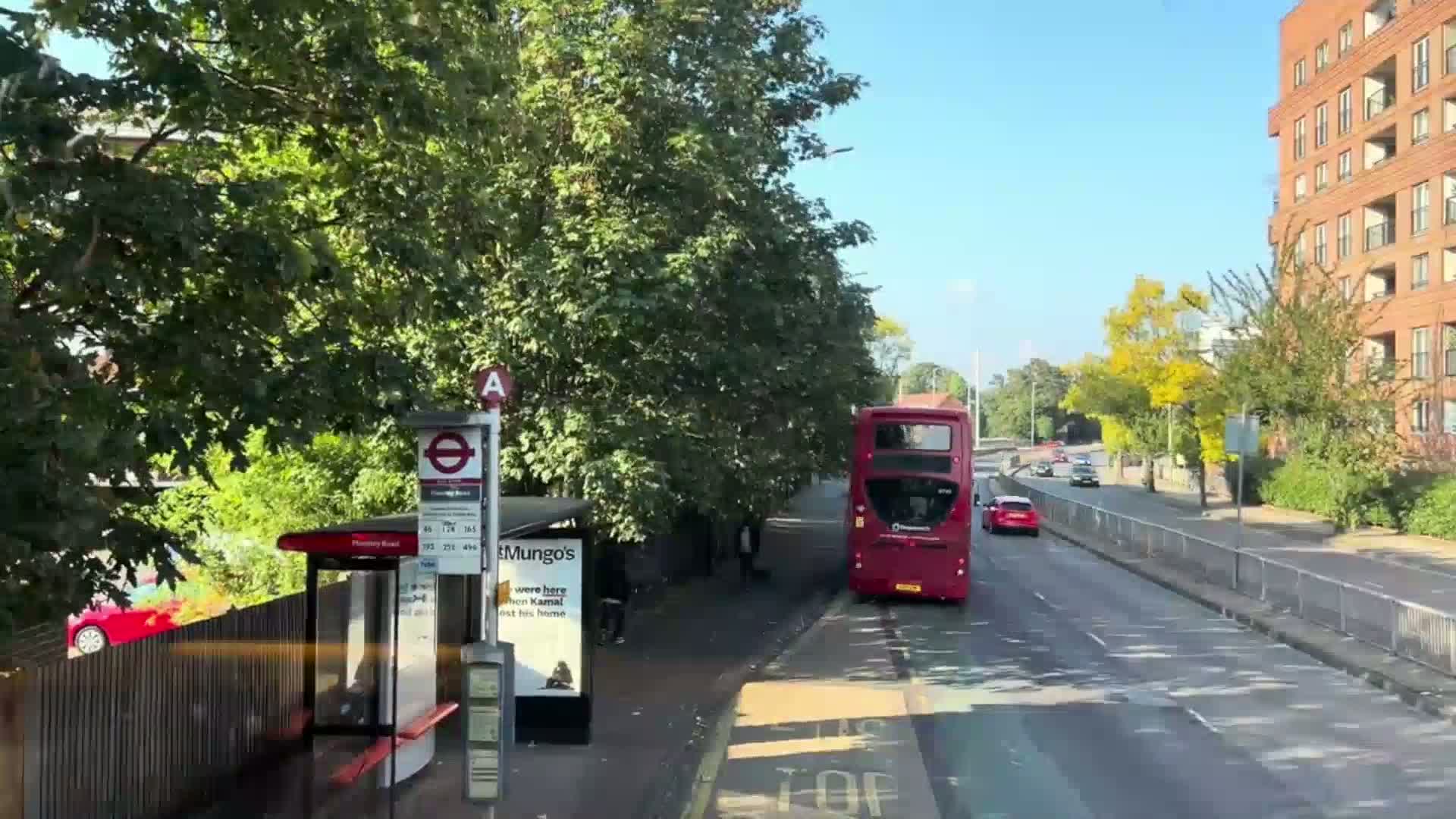 A red double-decker bus is stopped at a bus stop on a sunny day, next to a residential building and several trees.