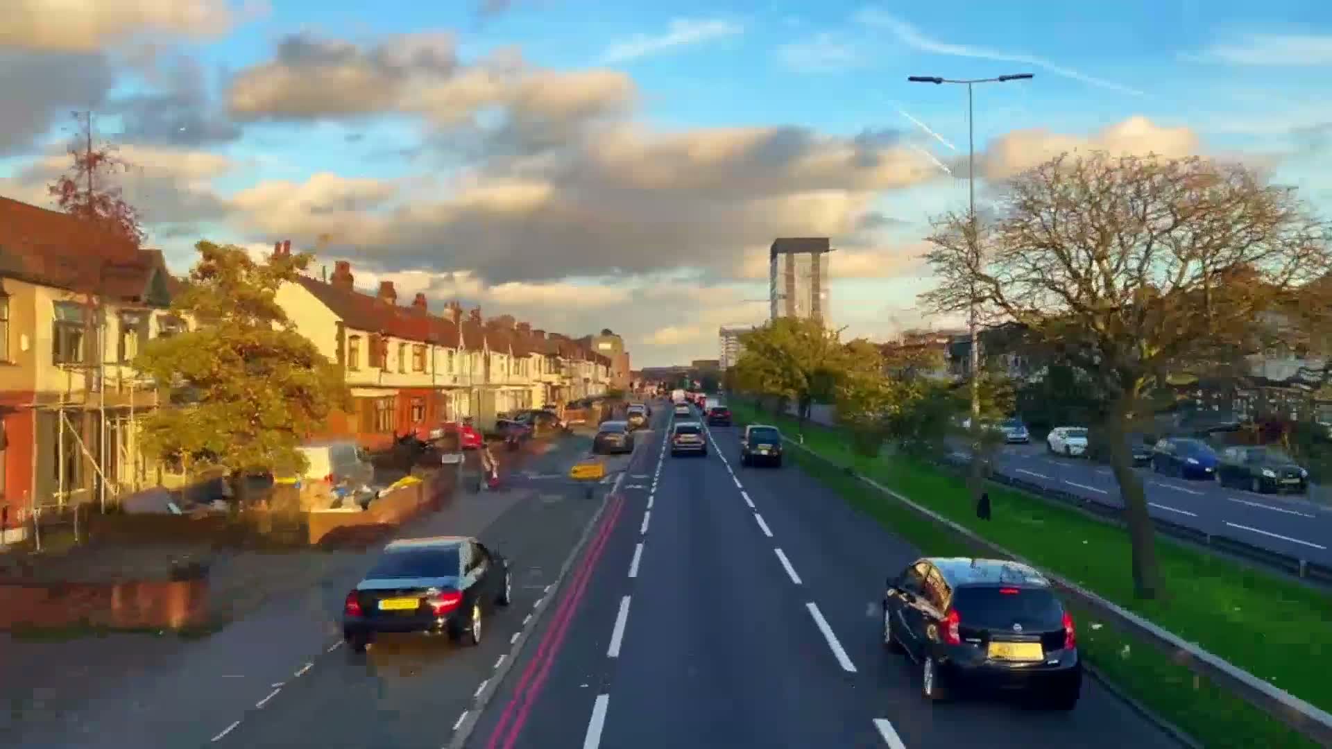 A partly cloudy, sunny day reveals a residential street lined with terraced houses, alongside a busy road with various cars driving in both directions.
