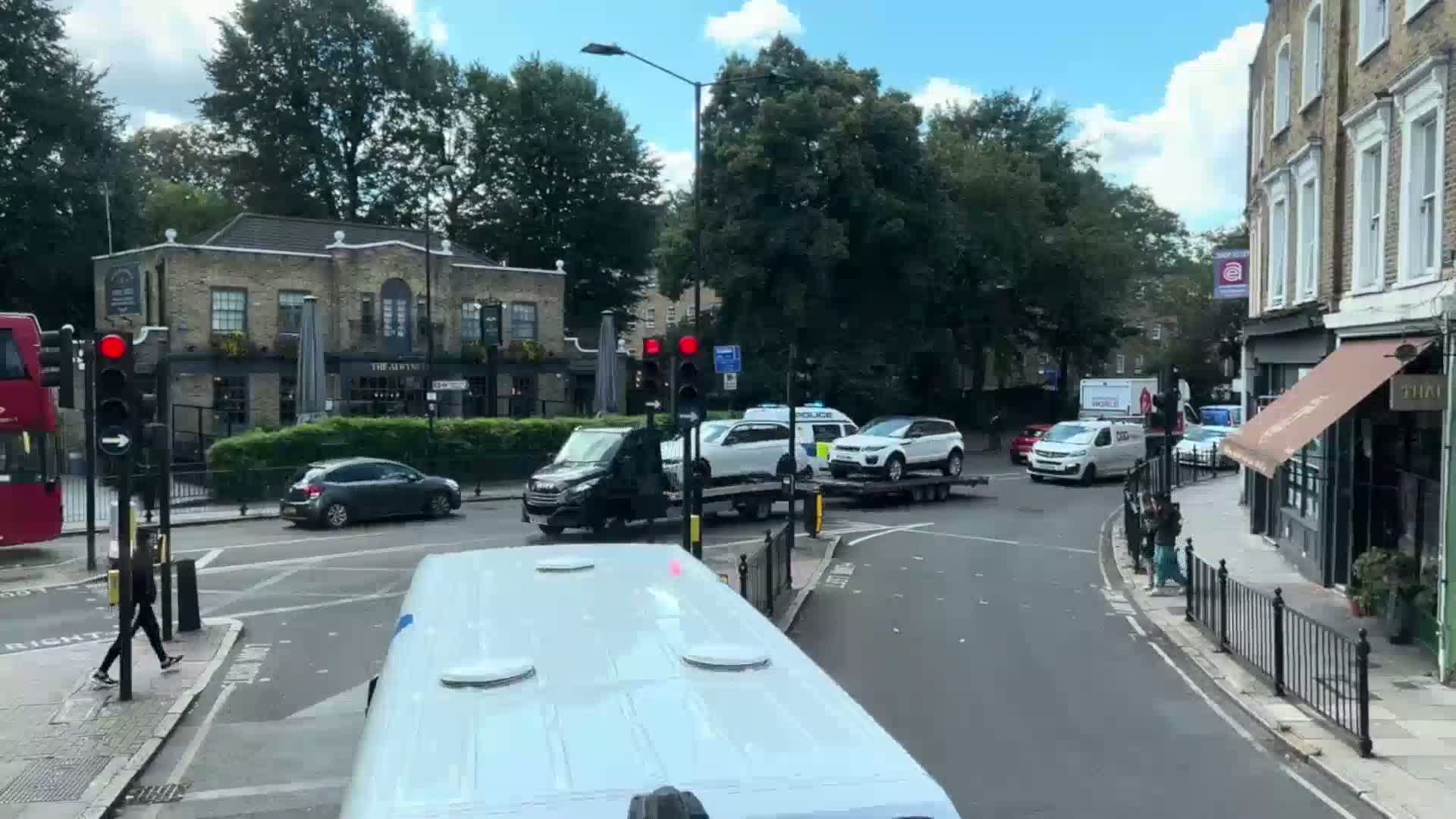 A flatbed tow truck carrying two cars is stopped at a city intersection on a sunny day, with pedestrians walking nearby.
