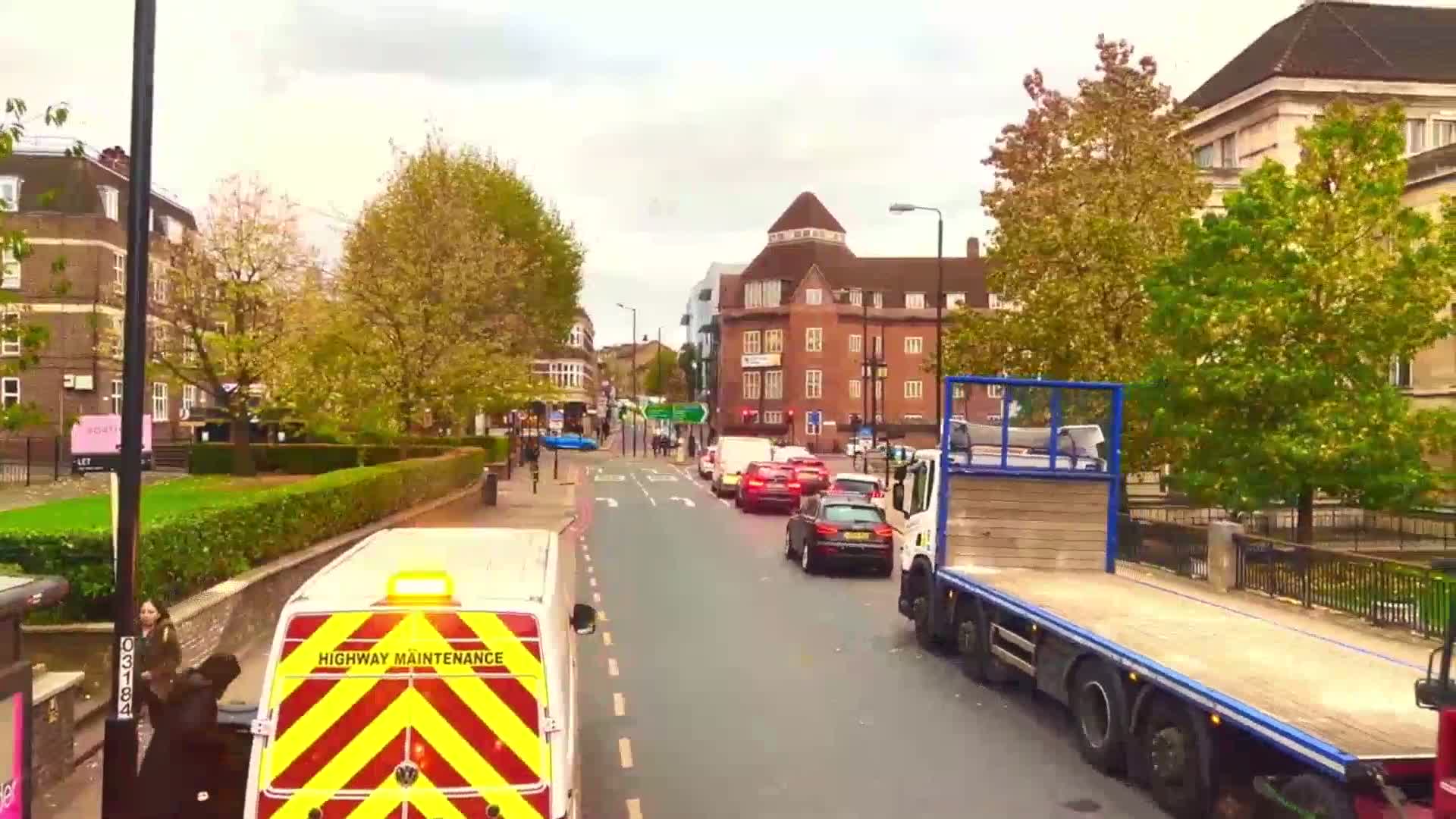 A highway maintenance van is driving down a city street with overcast skies, past other vehicles and buildings lined with trees beginning to change color.