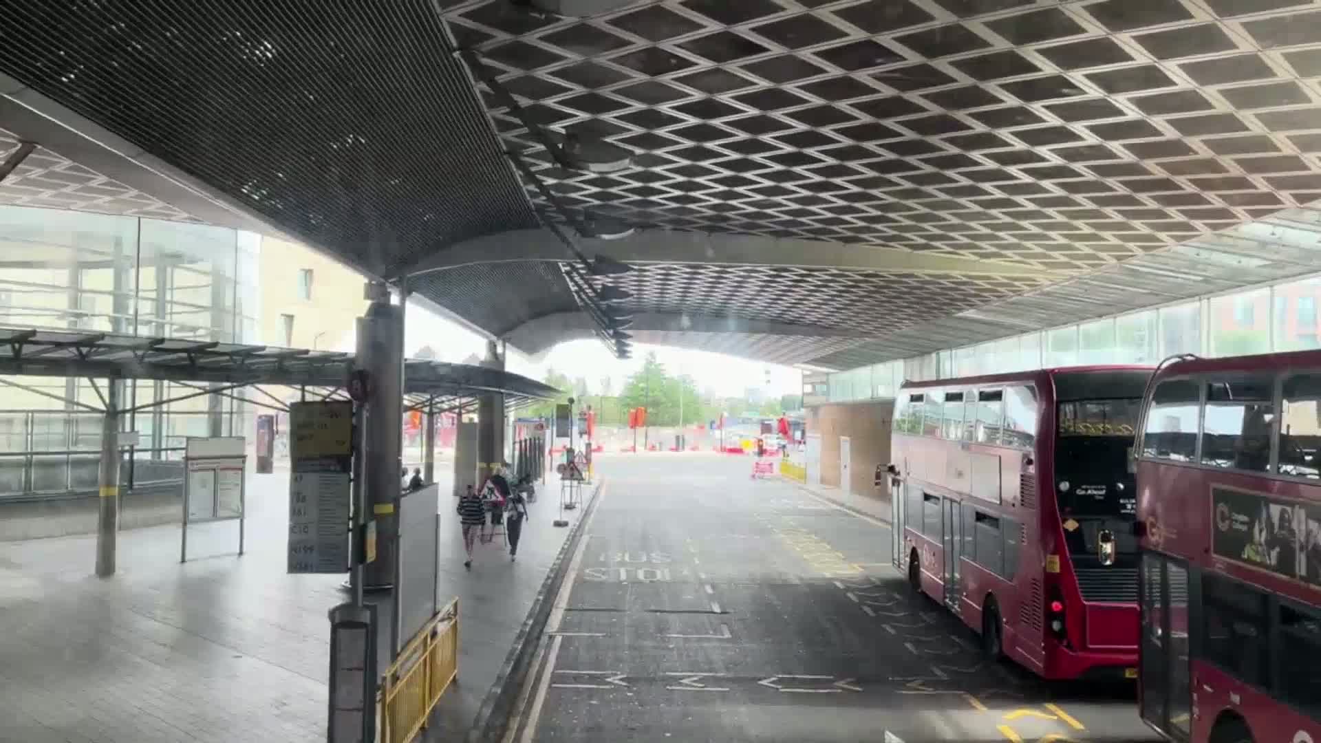 Under a covered bus stop on a partly sunny day, two double-decker buses are parked, and a few pedestrians are walking.