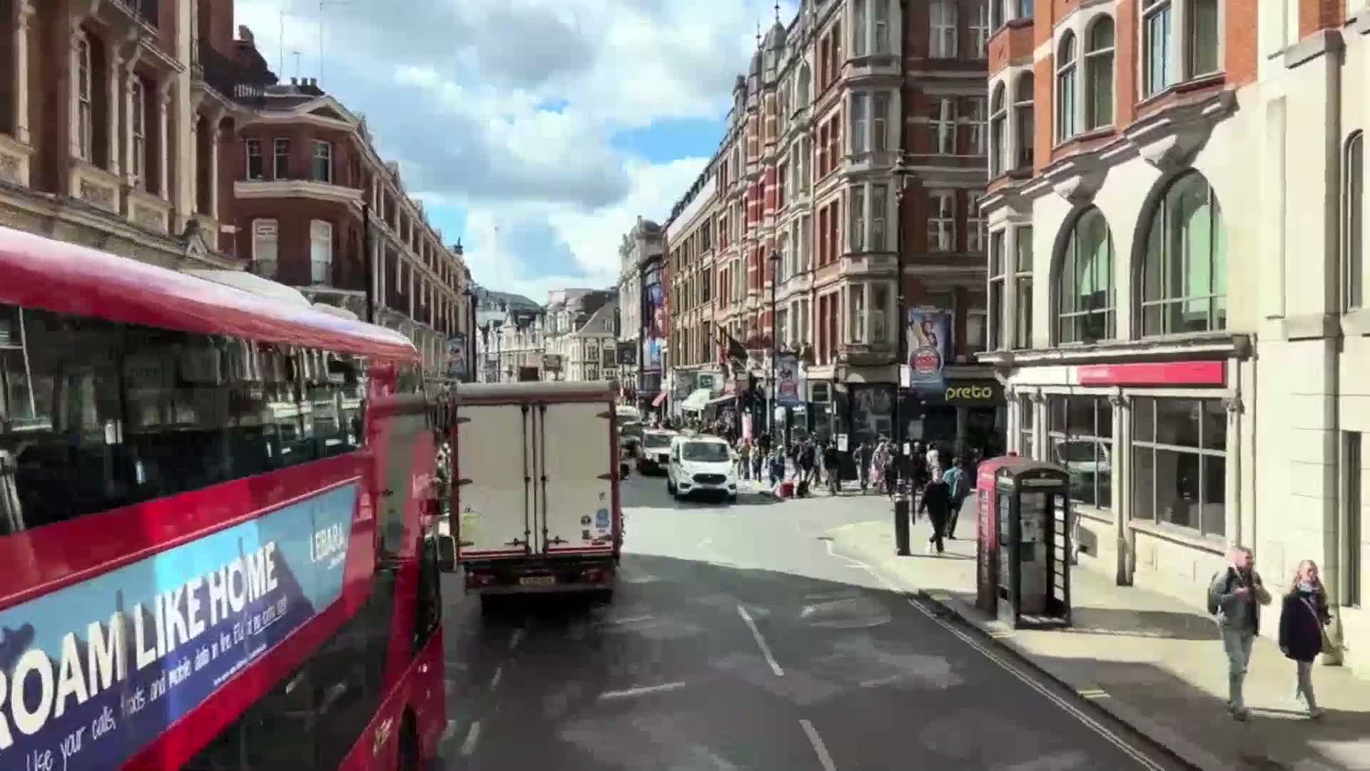 A red double-decker bus drives down a city street on a partly sunny day, passing other vehicles and pedestrians near multi-story buildings.