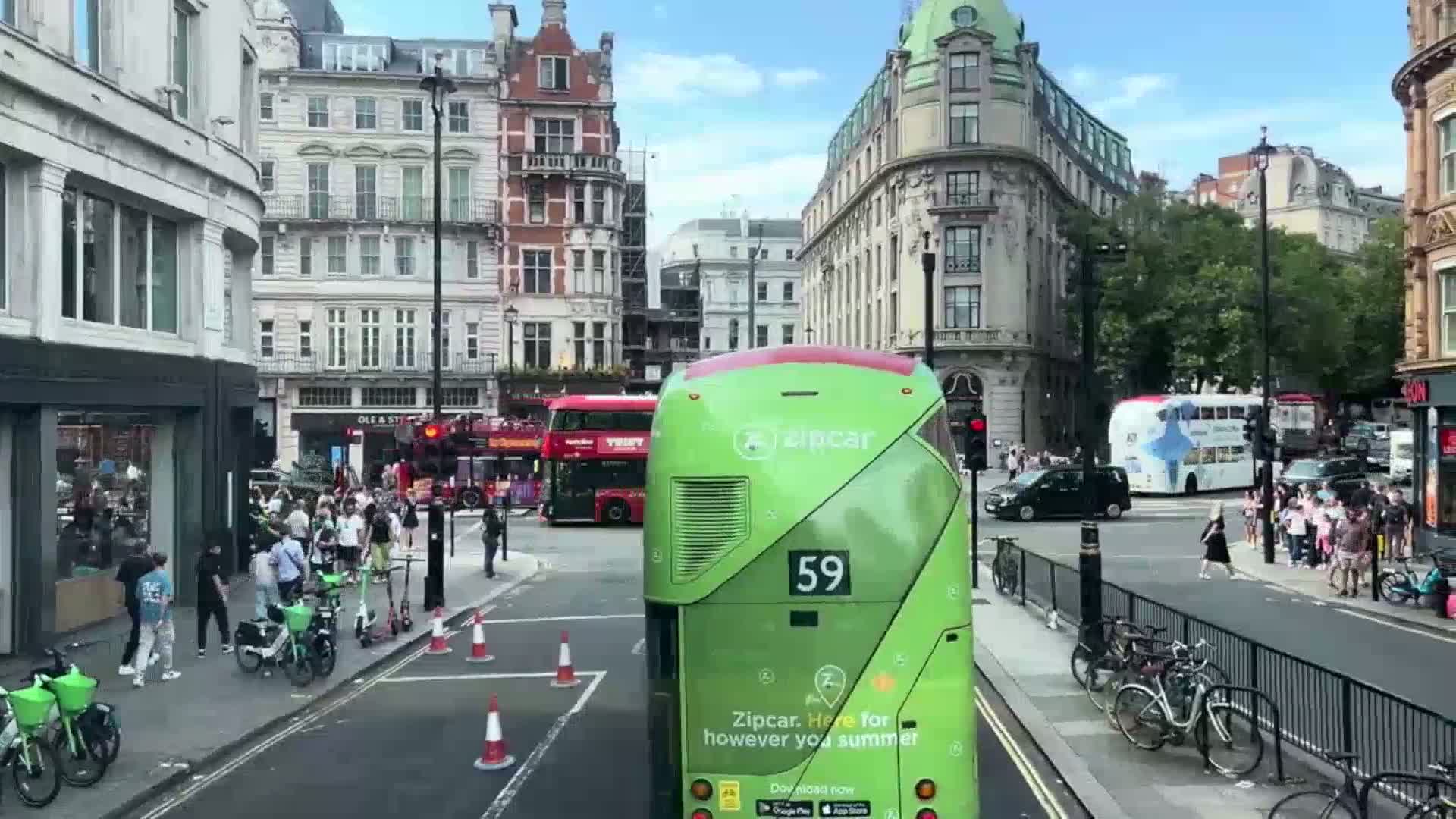 A green double-decker bus sits at an intersection in a city with several other buses, cars, pedestrians, and bicycles on a sunny day.