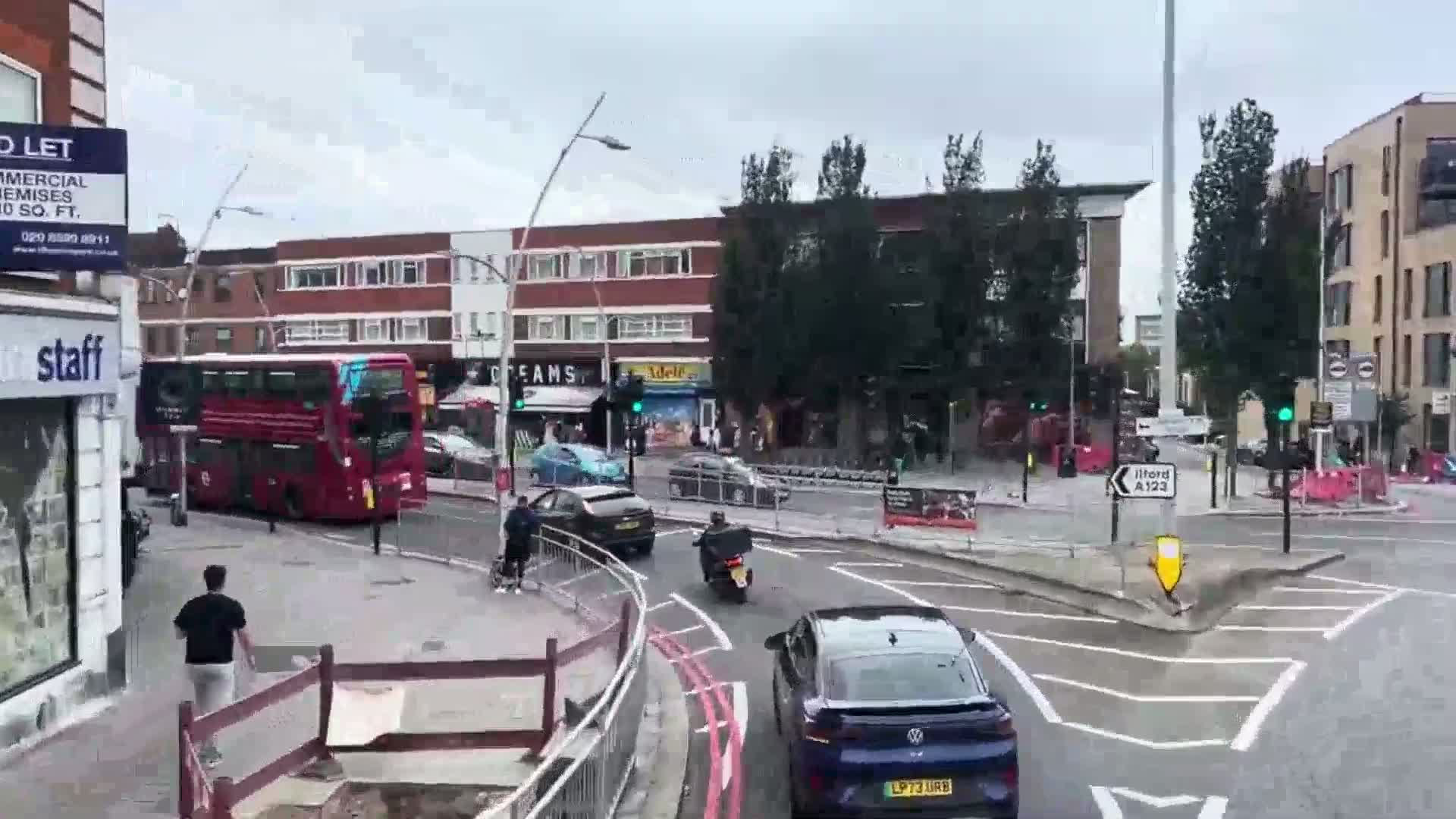 A cloudy day shows a busy intersection with a double-decker bus, several cars, a motor scooter, and pedestrians near shops and buildings under construction.