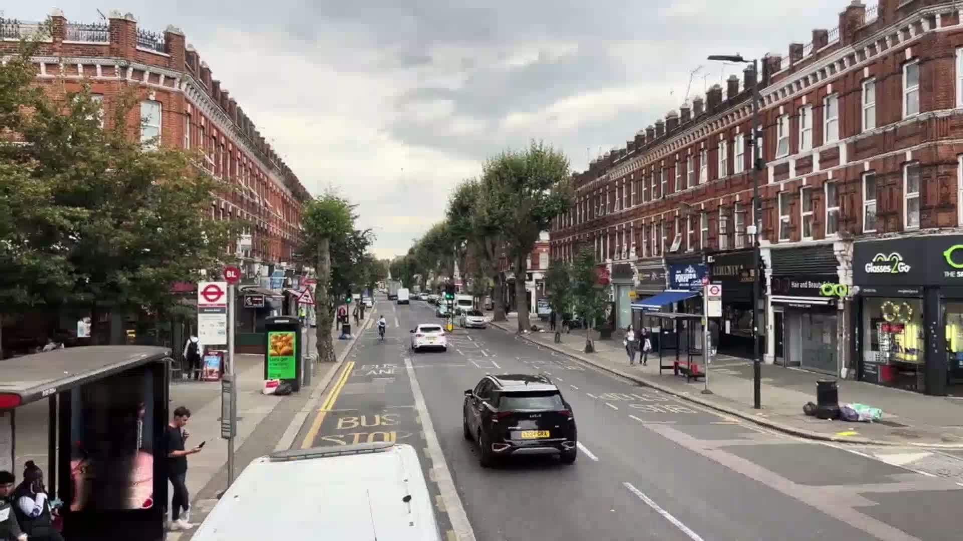 A street lined with brick buildings and trees under an overcast sky has cars and pedestrians on a partly sunny day.