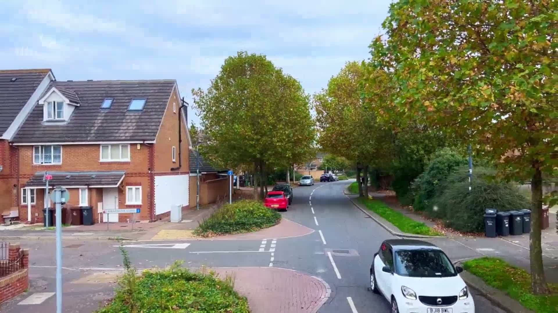 A residential street lined with brick houses and autumnal trees under a partly cloudy sky shows several parked cars and one driving a small white car.