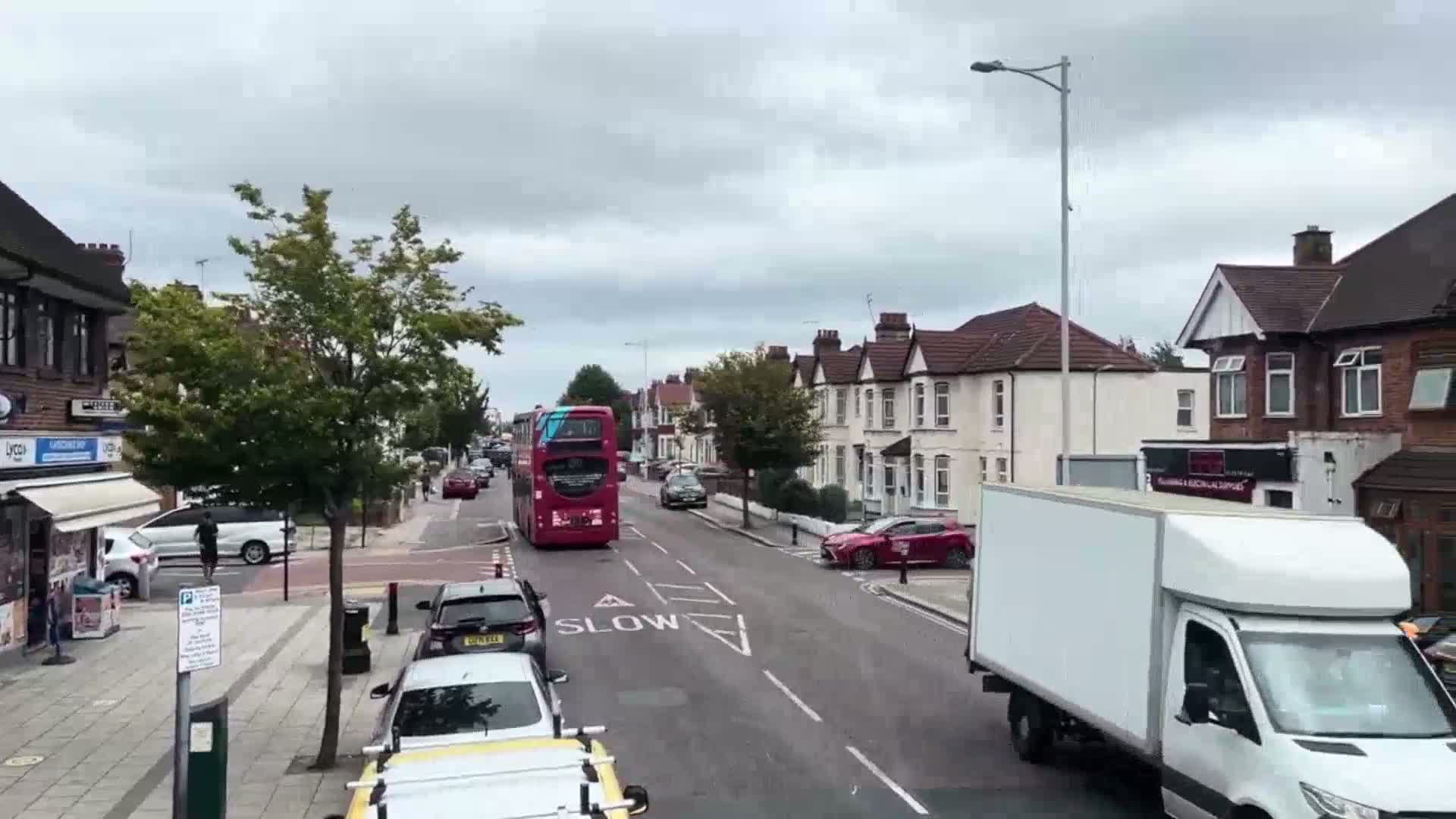 A red double-decker bus drives down a residential street on an overcast day,  past parked cars and houses with front gardens.