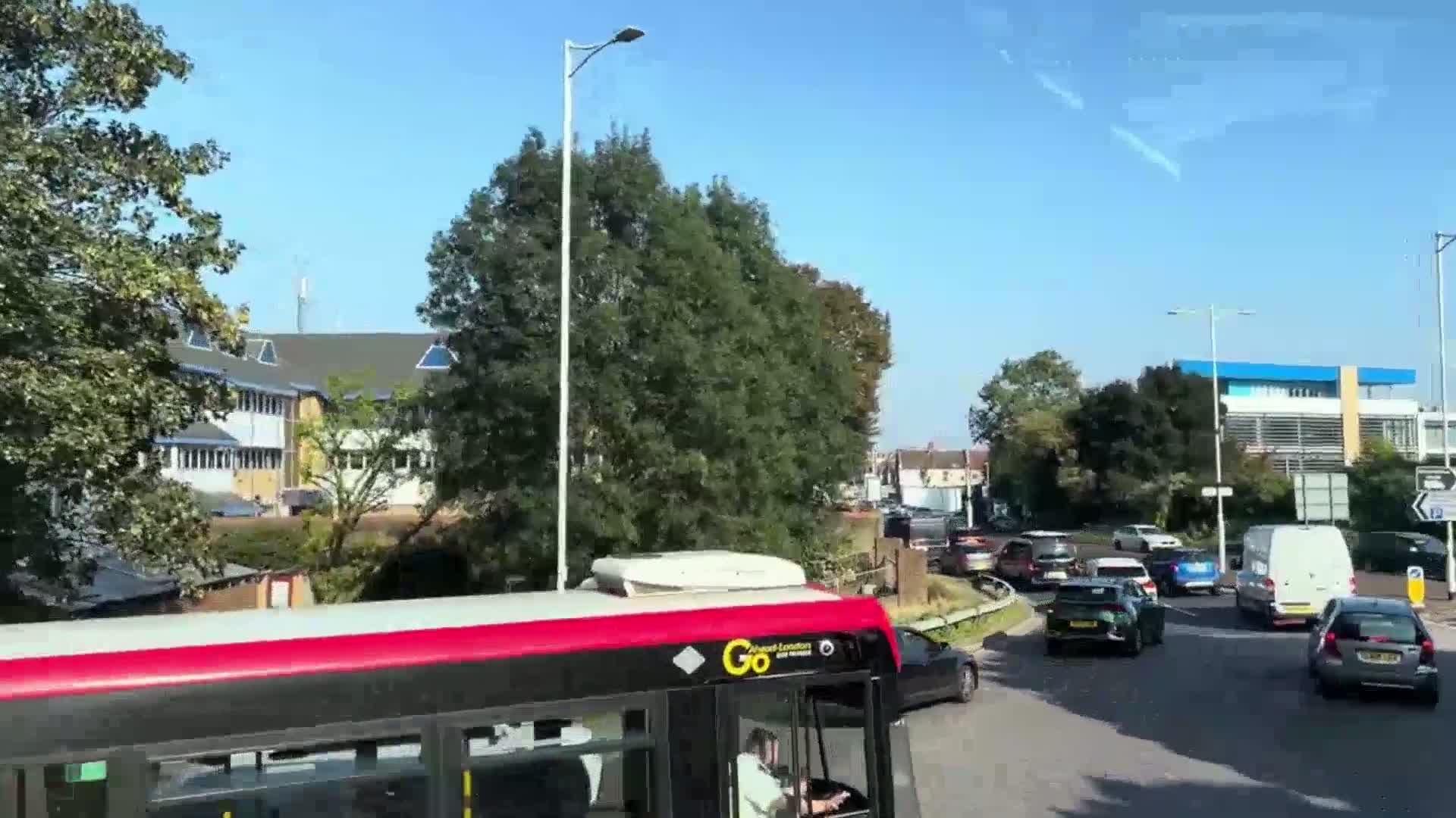 A red and black bus drives through a roundabout on a sunny day, surrounded by other vehicles and buildings.