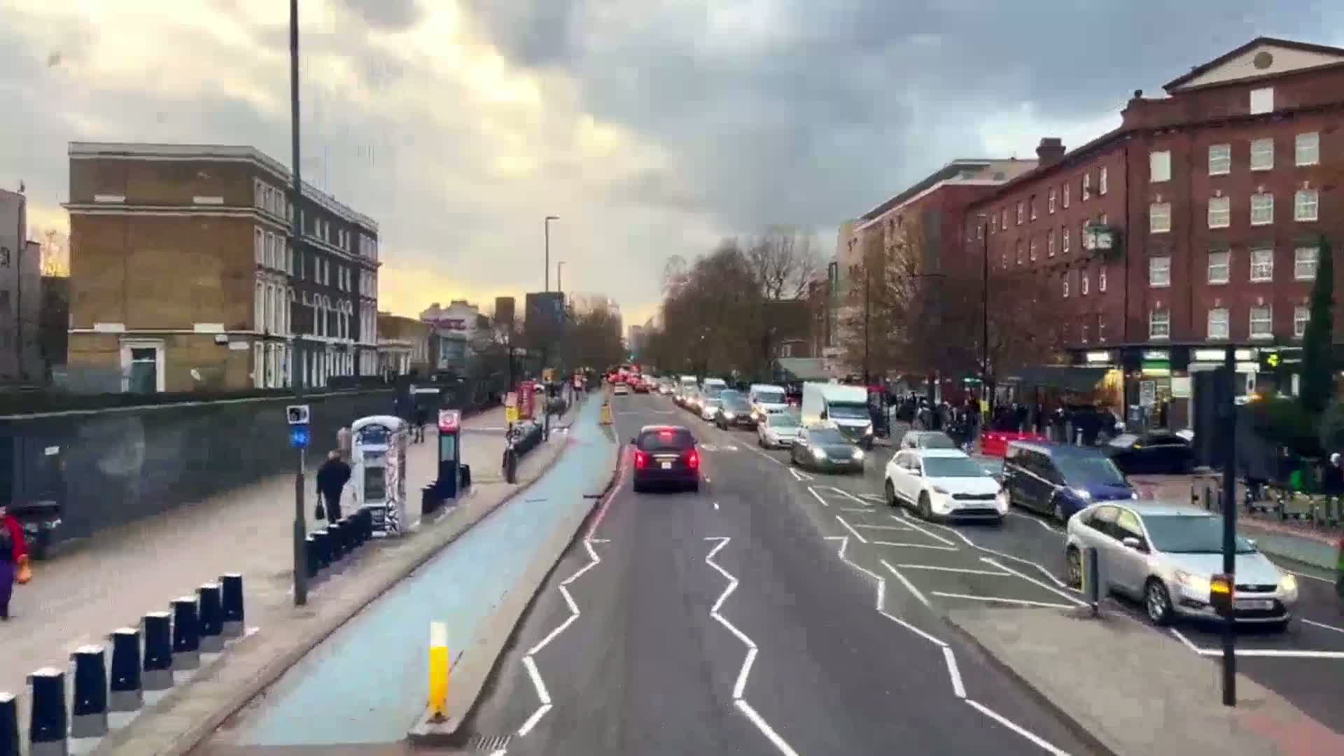 A city street scene under a cloudy sky shows several cars driving on a multi-lane road that has a dedicated bike lane and pedestrian areas next to buildings on both sides.