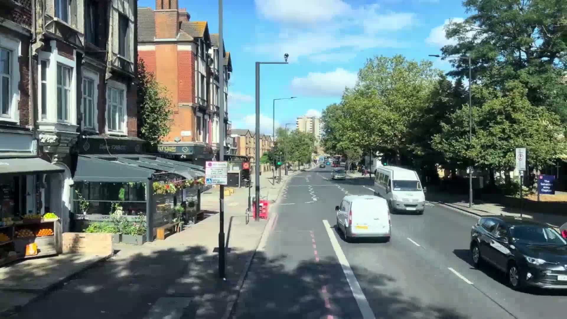 A sunny day on a city street with several cars and vans driving past brick buildings with shops and awnings on the side of the road.