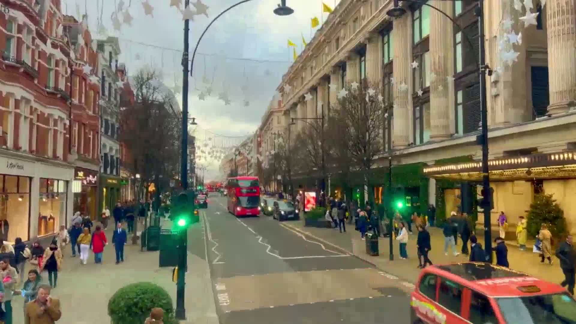 A city street lined with shops and pedestrians, decorated with hanging star shapes, features a double-decker bus and other vehicles under an overcast sky.