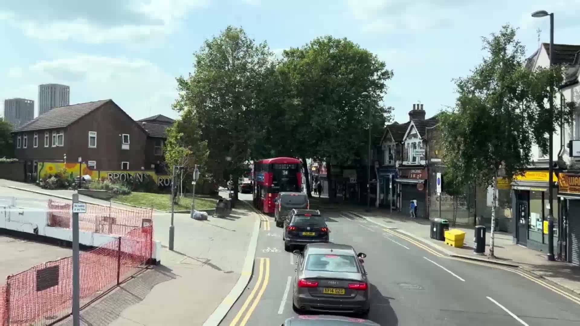 A partly sunny day shows a residential street with a double-decker bus and several cars, some cyclists, and pedestrians near shops and buildings.