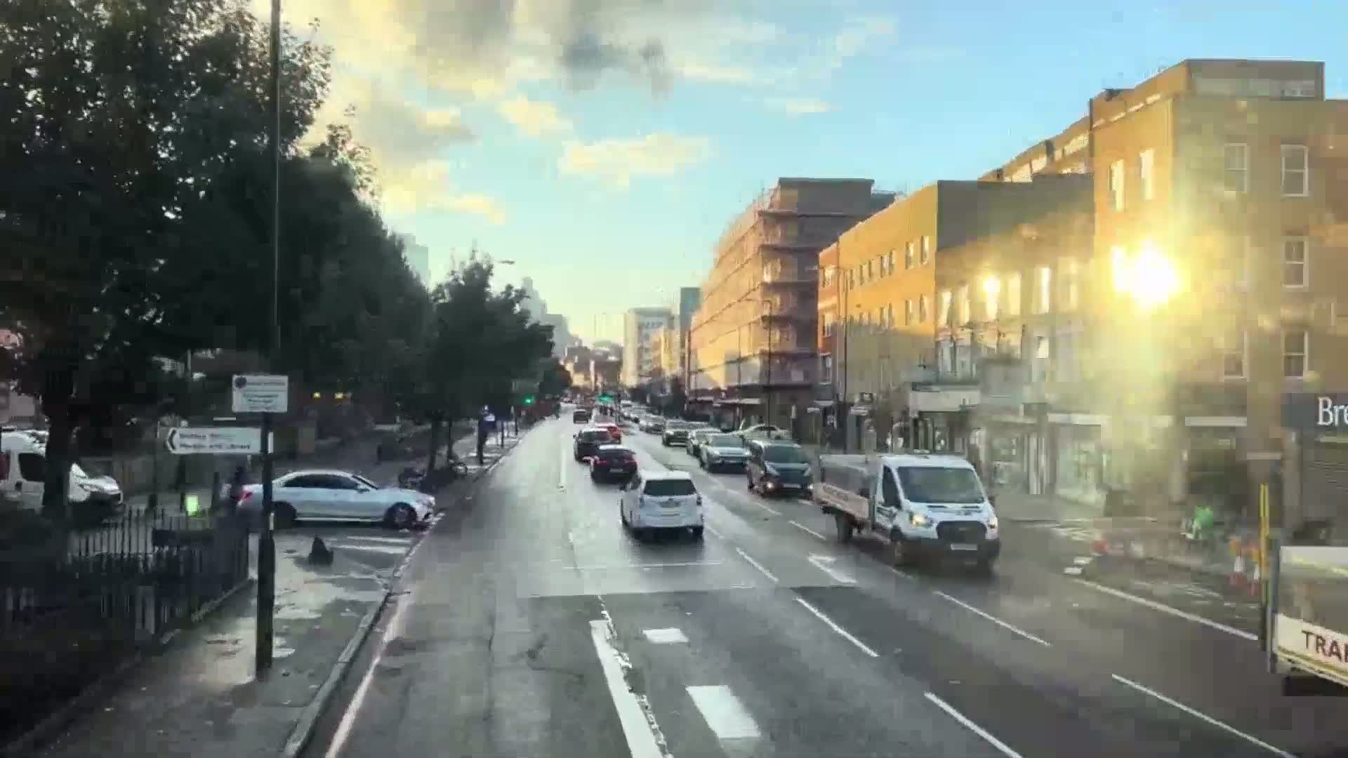 A wet city street with several cars driving in both directions, lined with multi-story buildings under a partly sunny sky.