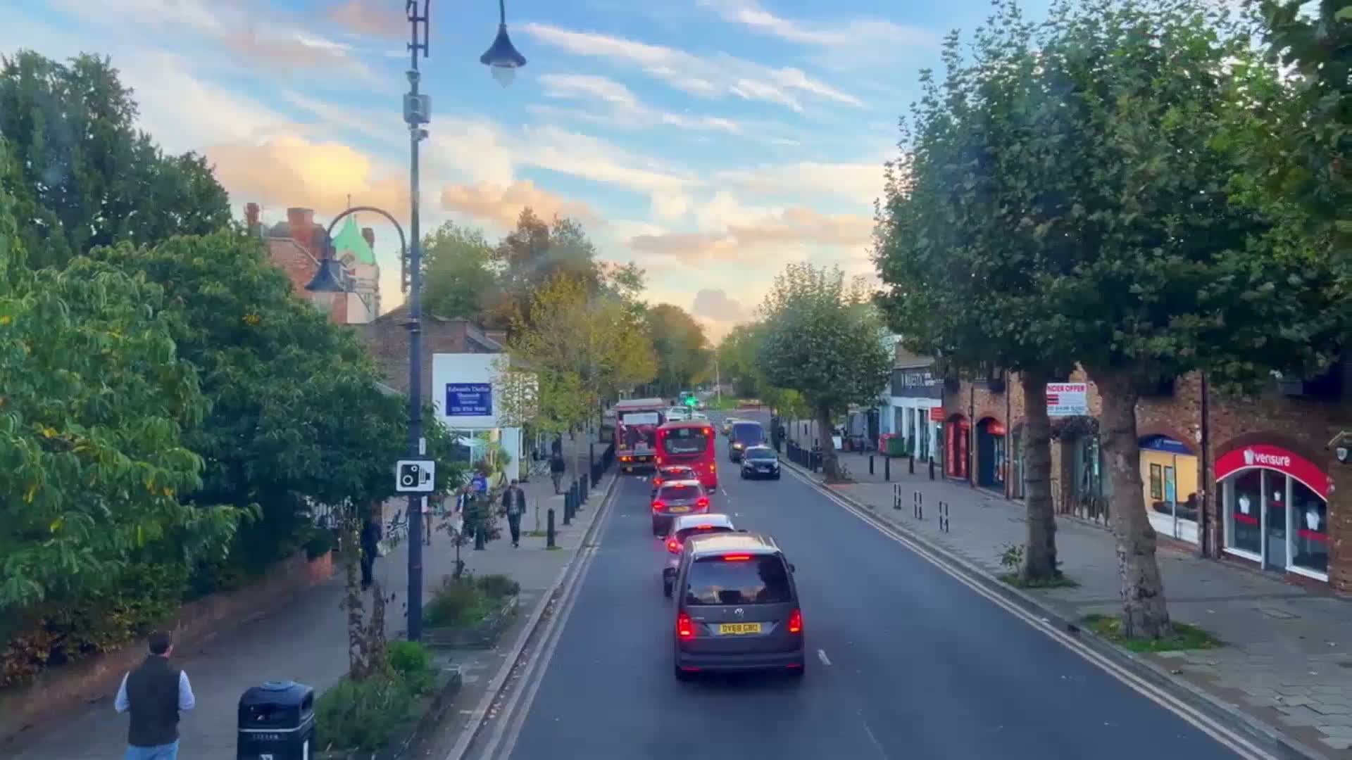 A partly cloudy afternoon street scene shows a line of cars and two double-decker buses stopped in traffic, with pedestrians walking on the sidewalk and shops lining the street.