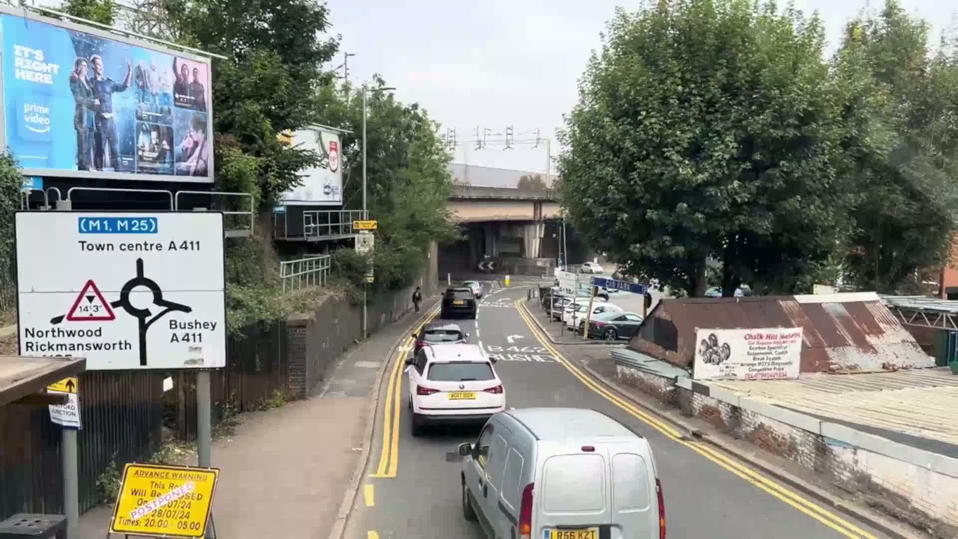 A road scene under an overcast sky shows several cars and a van traveling beneath a bridge, with trees and buildings lining the street.