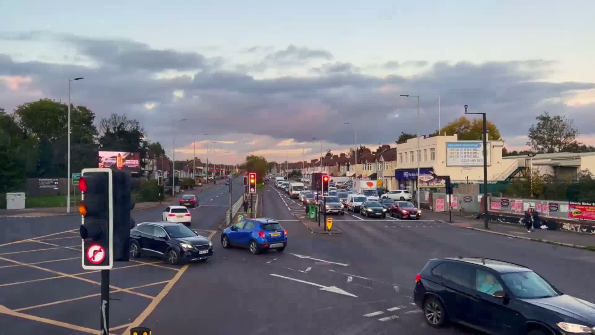 A multi-lane road intersection with moderate traffic under a partly cloudy sky shows cars stopped at traffic lights.
