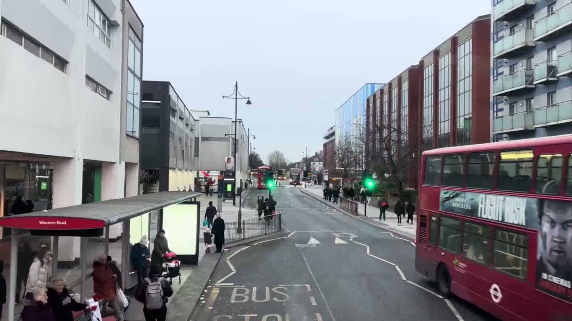 A city street scene on an overcast day shows a double-decker bus driving past a bus stop where several people are waiting, with more pedestrians walking along the sidewalk and another bus further down the street.