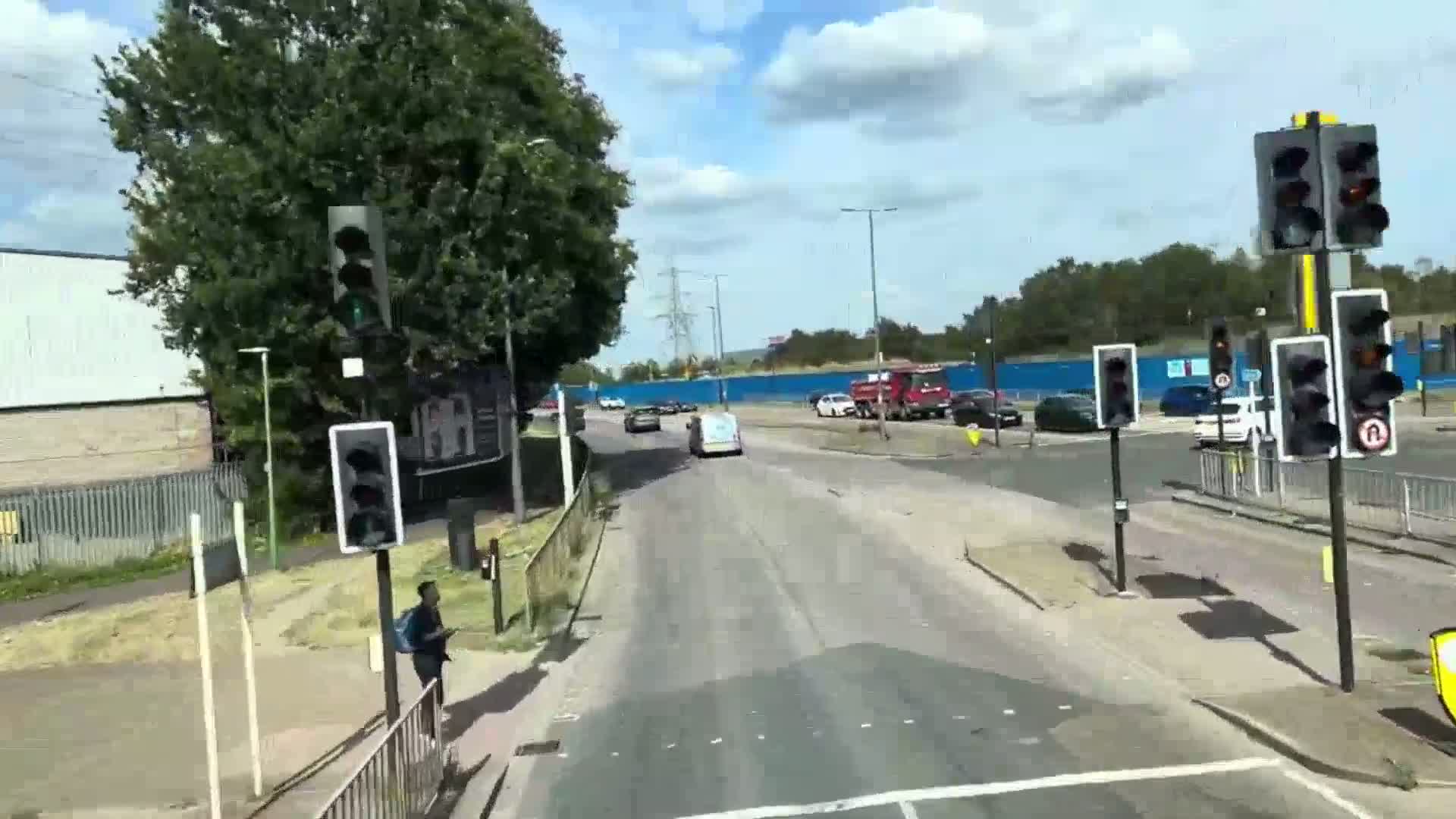 A partly sunny day shows a road intersection with several cars and a person walking near traffic lights; a large blue construction barrier is visible in the background.