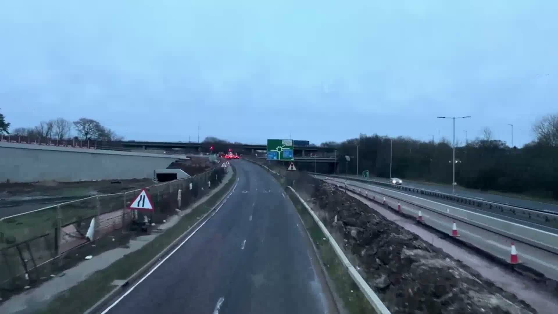 A road under construction curves gently to the right, passing under an overpass, with a few cars visible on both the main road and the adjacent highway on an overcast day.