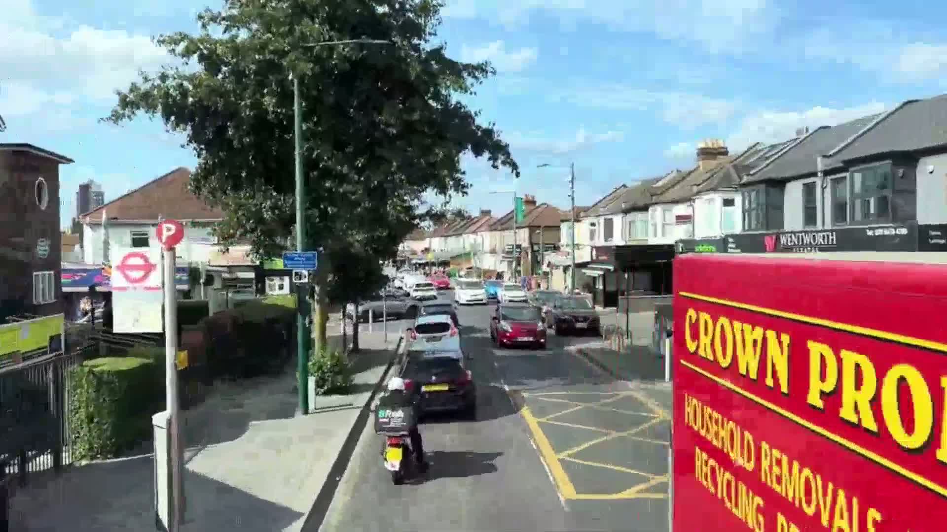 A sunny day reveals a residential street with parked and moving cars, a motorcyclist, and a row of shops along the roadside.
