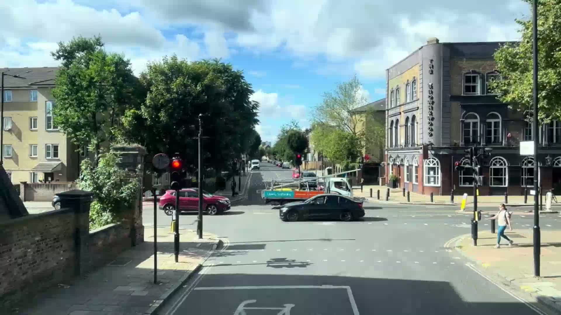 A sunny day shows a city intersection with cars at a traffic light, pedestrians crossing the street, and buildings lining the road.