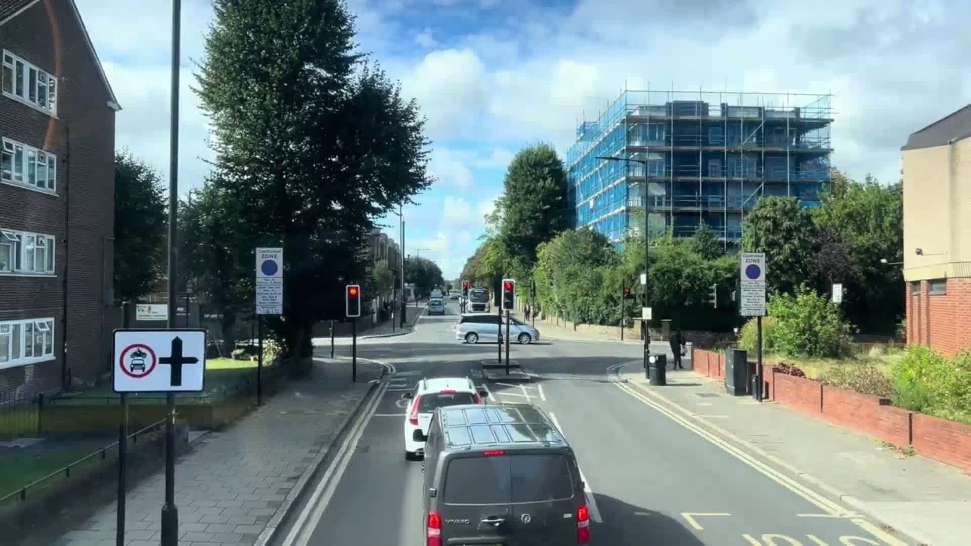 A partly sunny day shows a residential street with several cars at a traffic intersection, a building under construction, and lush green trees.