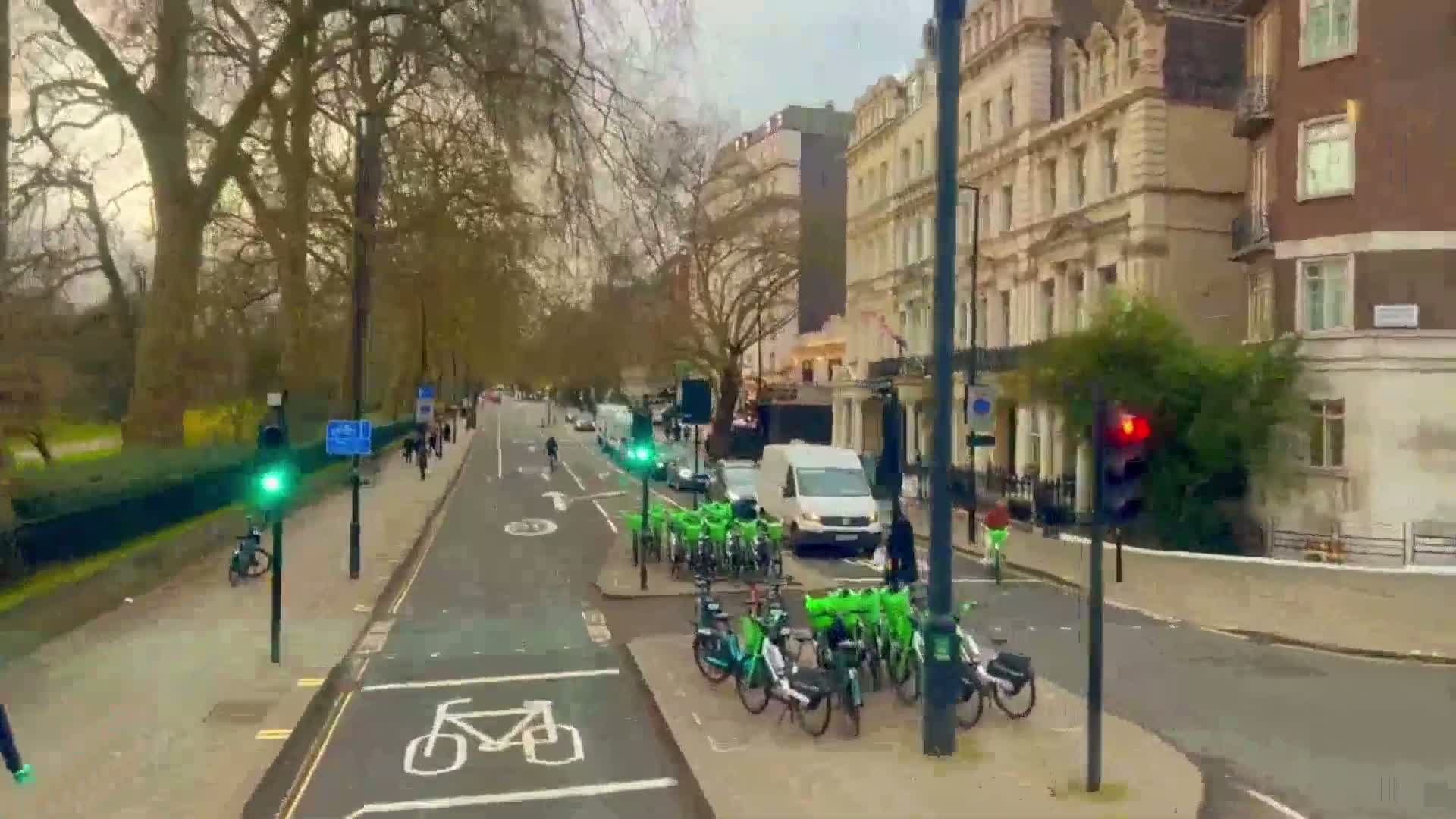 A city street scene on an overcast day shows a row of lime green rental bikes parked near a traffic light, with cars and pedestrians visible.