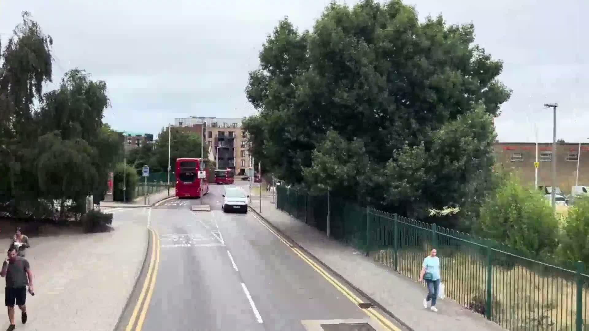 A street scene on an overcast day shows two red double-decker buses, a white van, and several pedestrians near a green fence and some apartment buildings.