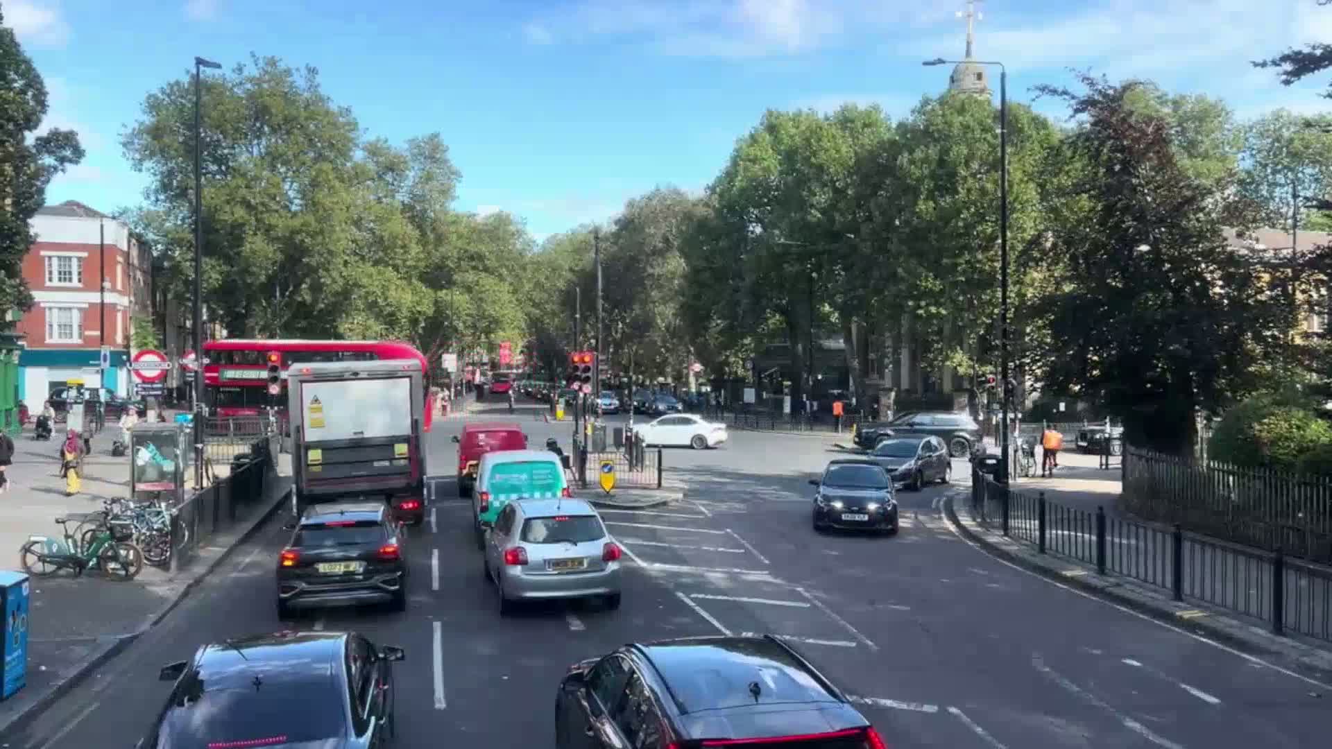 A sunny day shows a city street intersection with several cars, a bus, and a delivery truck at a standstill, surrounded by lush green trees and buildings.