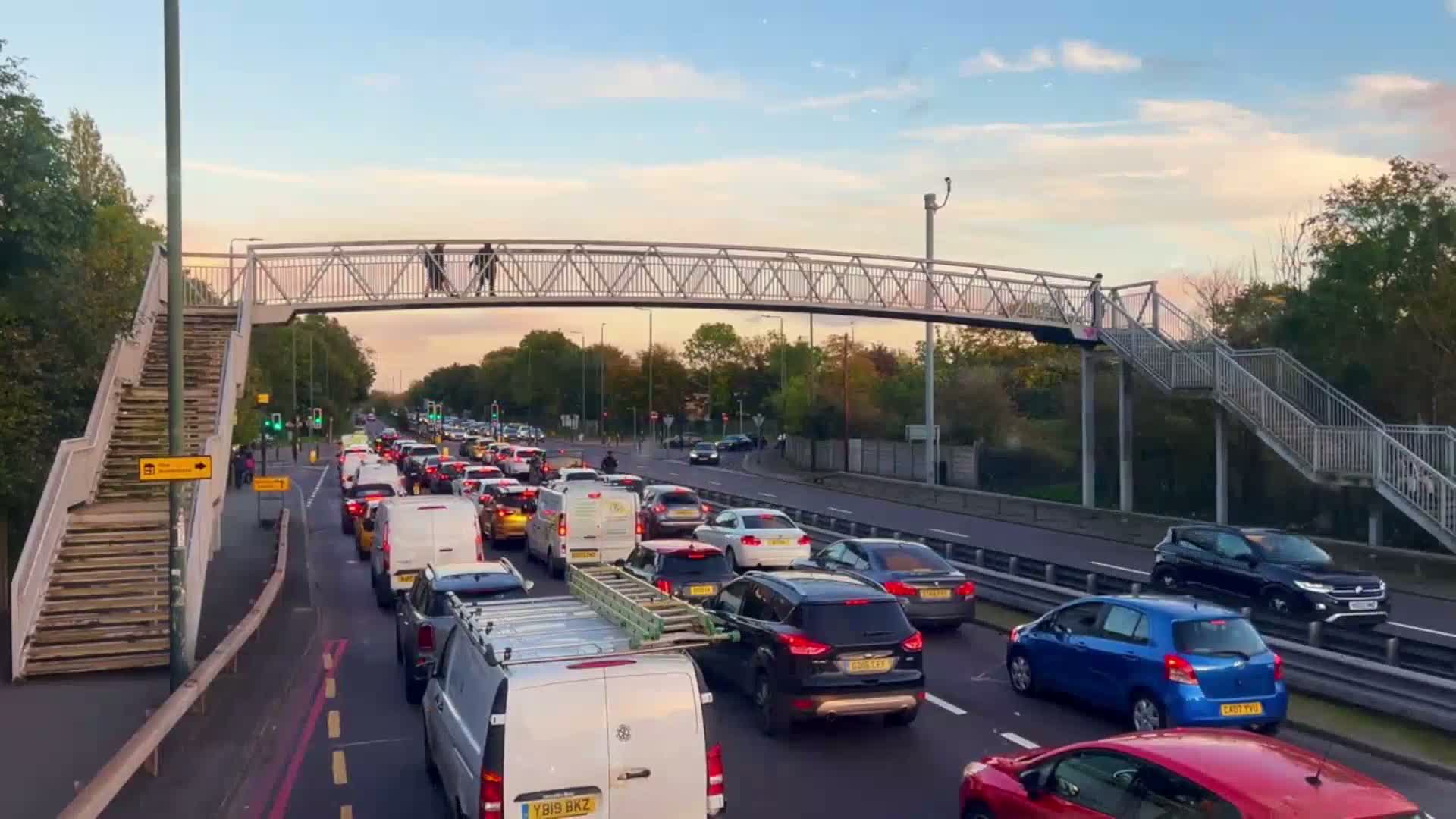 A pedestrian bridge spans a busy road with heavy traffic under a partly cloudy, twilight sky.