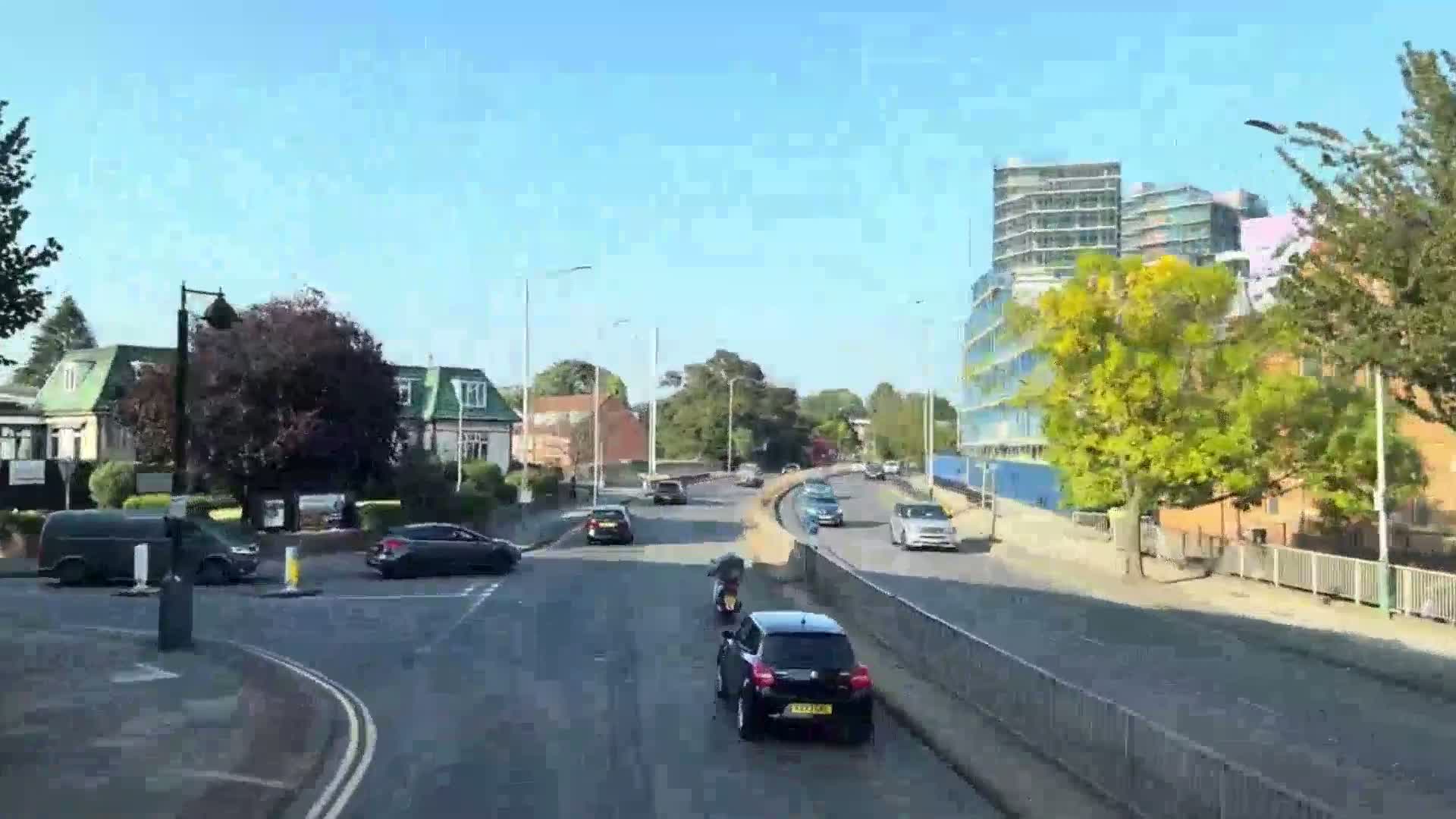 A sunny day reveals a multi-lane road with several cars and a motorcyclist, flanked by residential buildings and a modern high-rise structure.