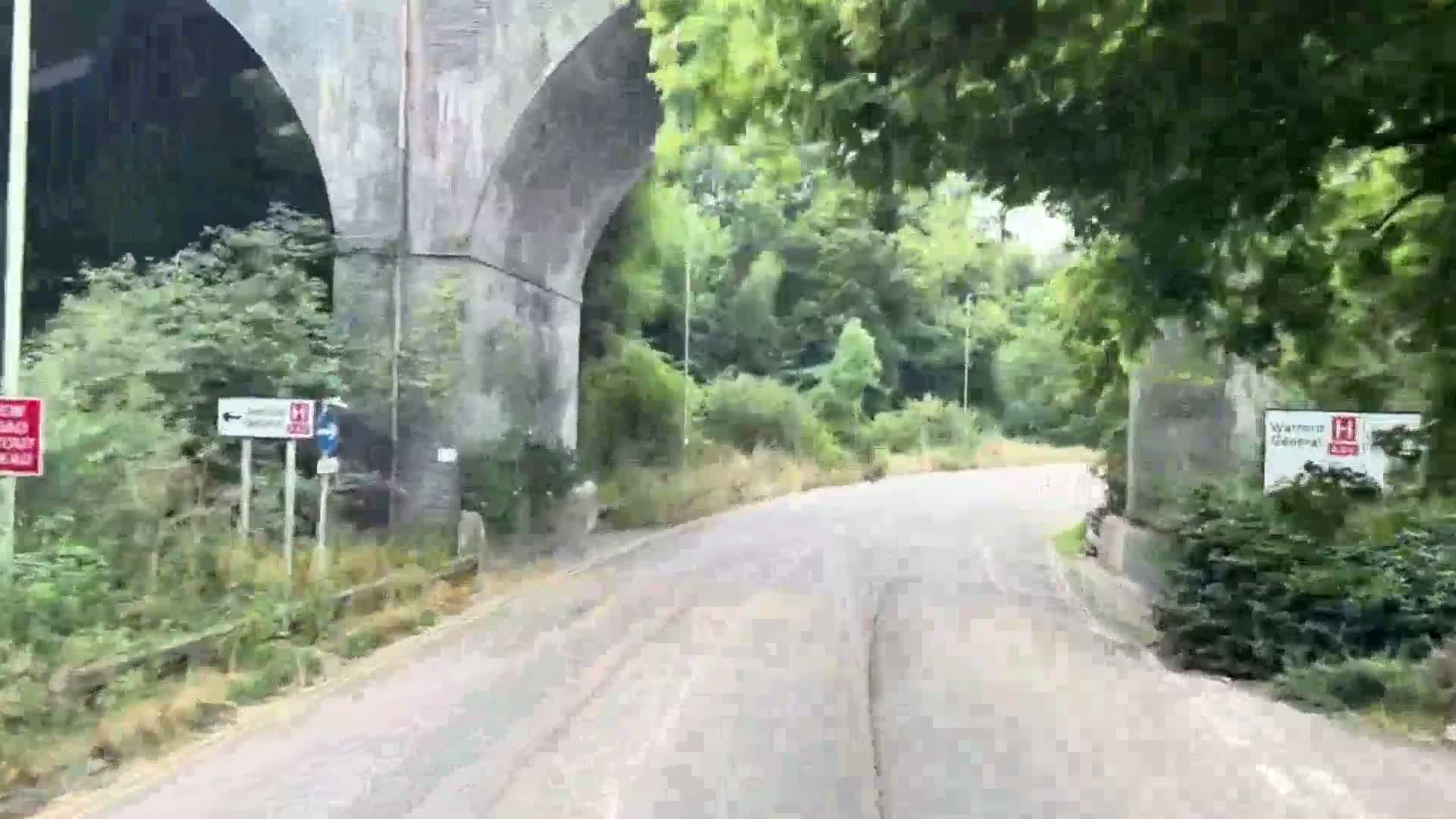 A paved road curves gently beneath a stone archway, passing through lush green vegetation under a partly sunny sky.