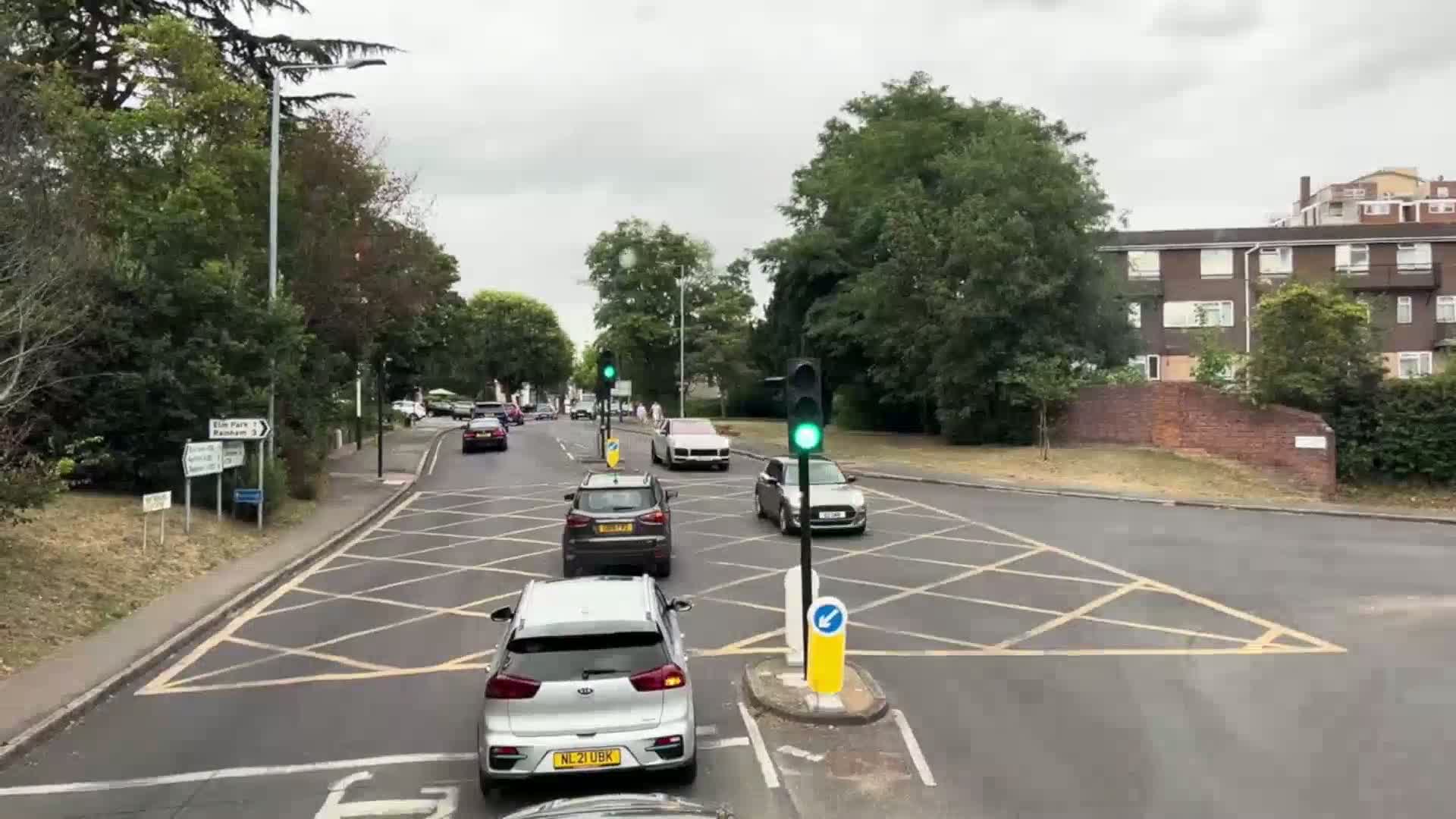 Several cars are navigating a crossroads under overcast skies, with lush green trees lining the road.