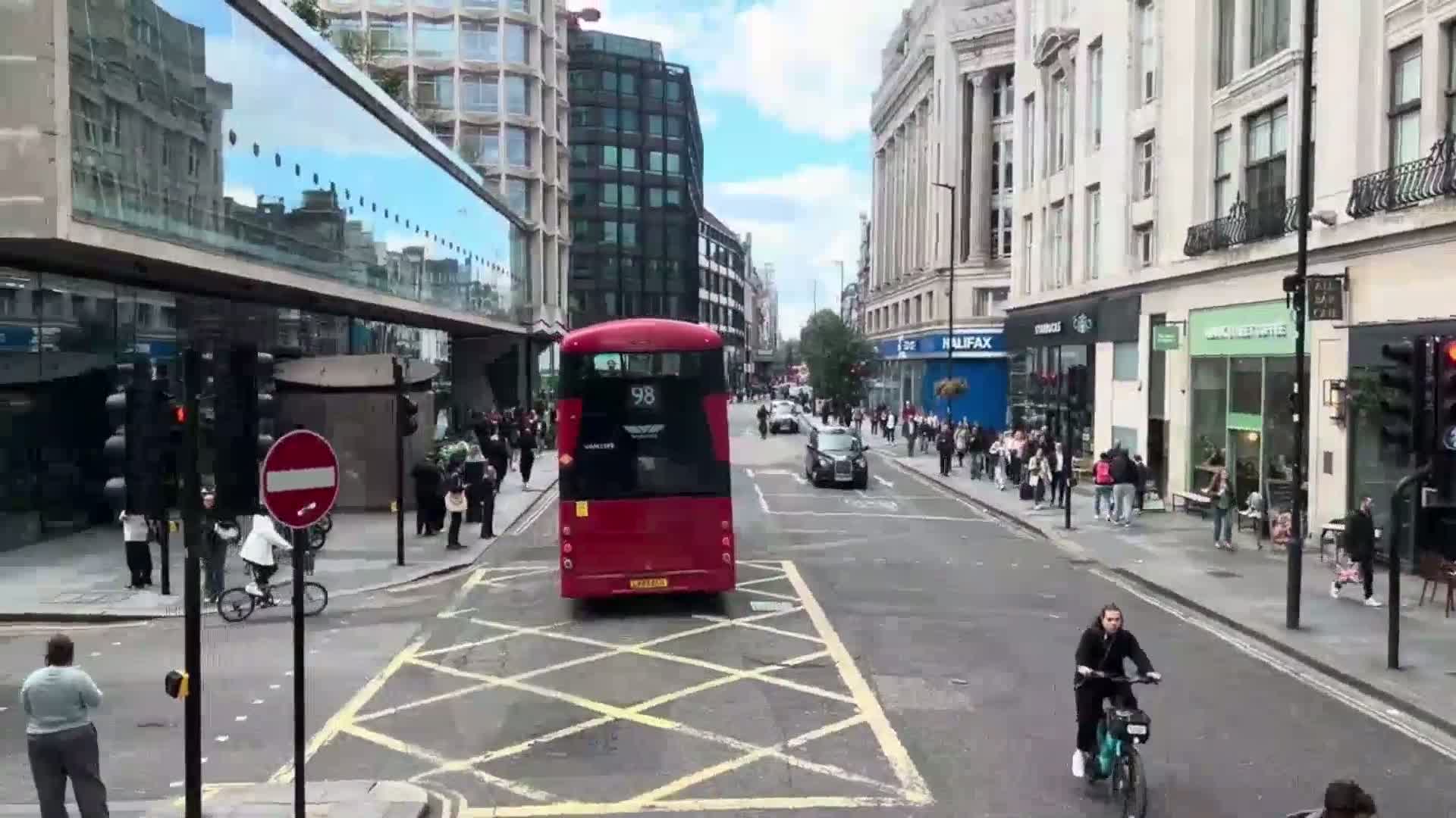 A red double-decker bus is stopped in the middle of a city street on a partly sunny day, with pedestrians and cyclists on the sidewalks and in the street.