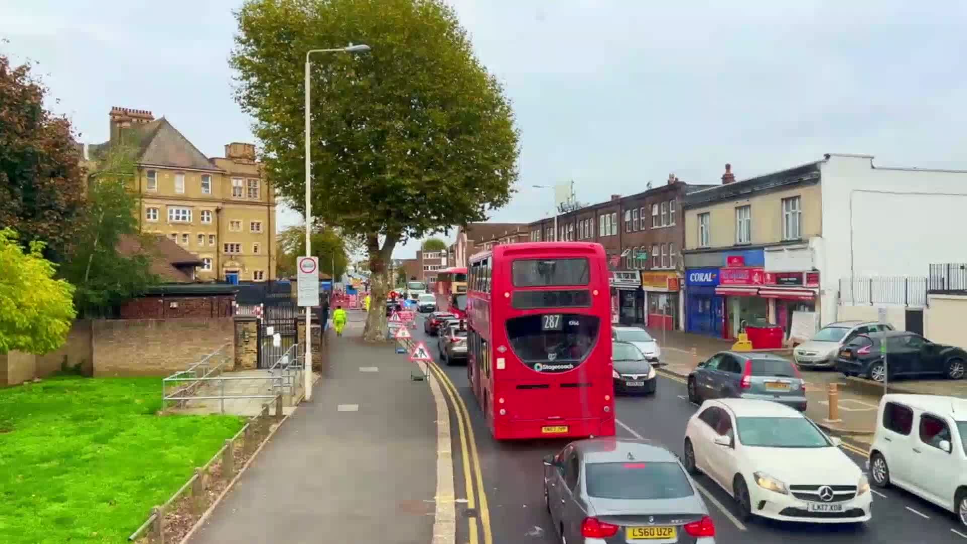 A red double-decker bus leads a line of cars down a street lined with shops and buildings under an overcast sky.