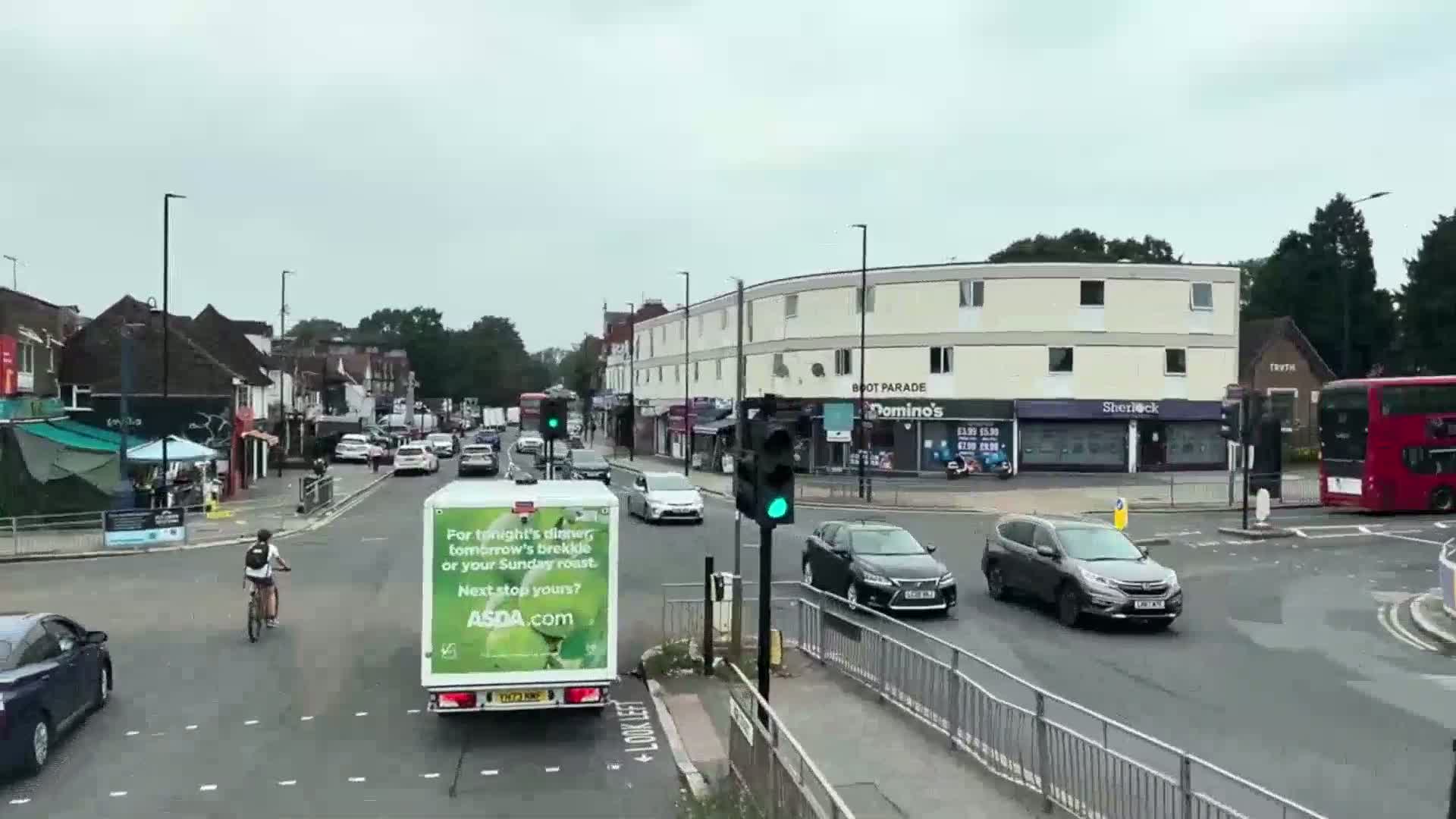 A cyclist rides past a busy intersection on an overcast day, with several cars and a large delivery truck at a traffic light, and a double-decker bus in the background.
