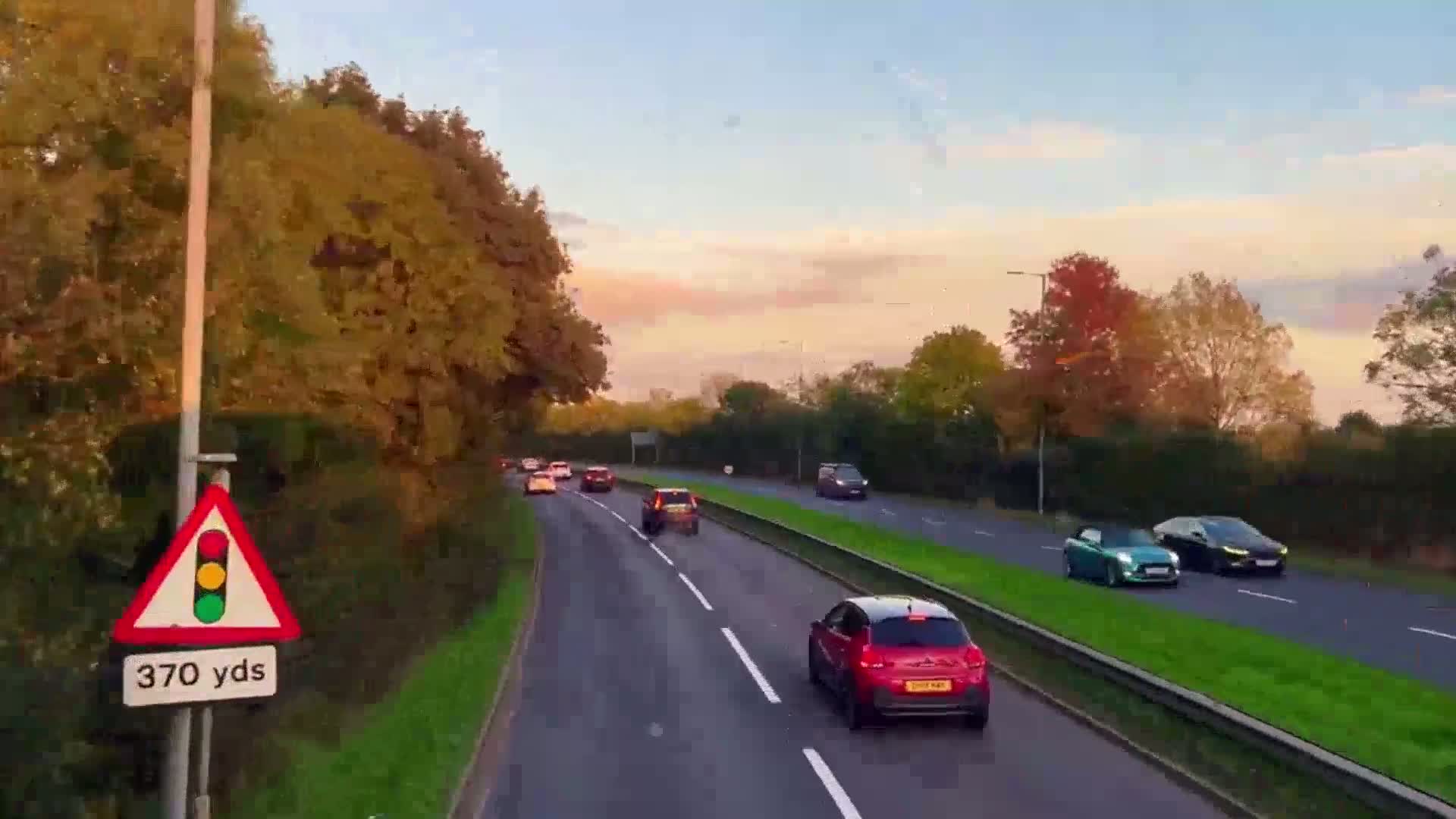 A multi-lane road with several cars driving in both directions under a partly cloudy, sunset sky, lined with autumnal trees.