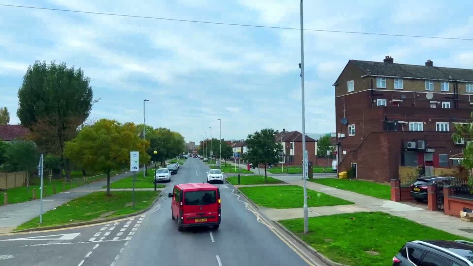 A red van drives down a residential street lined with trees and brick buildings under a partly cloudy sky.