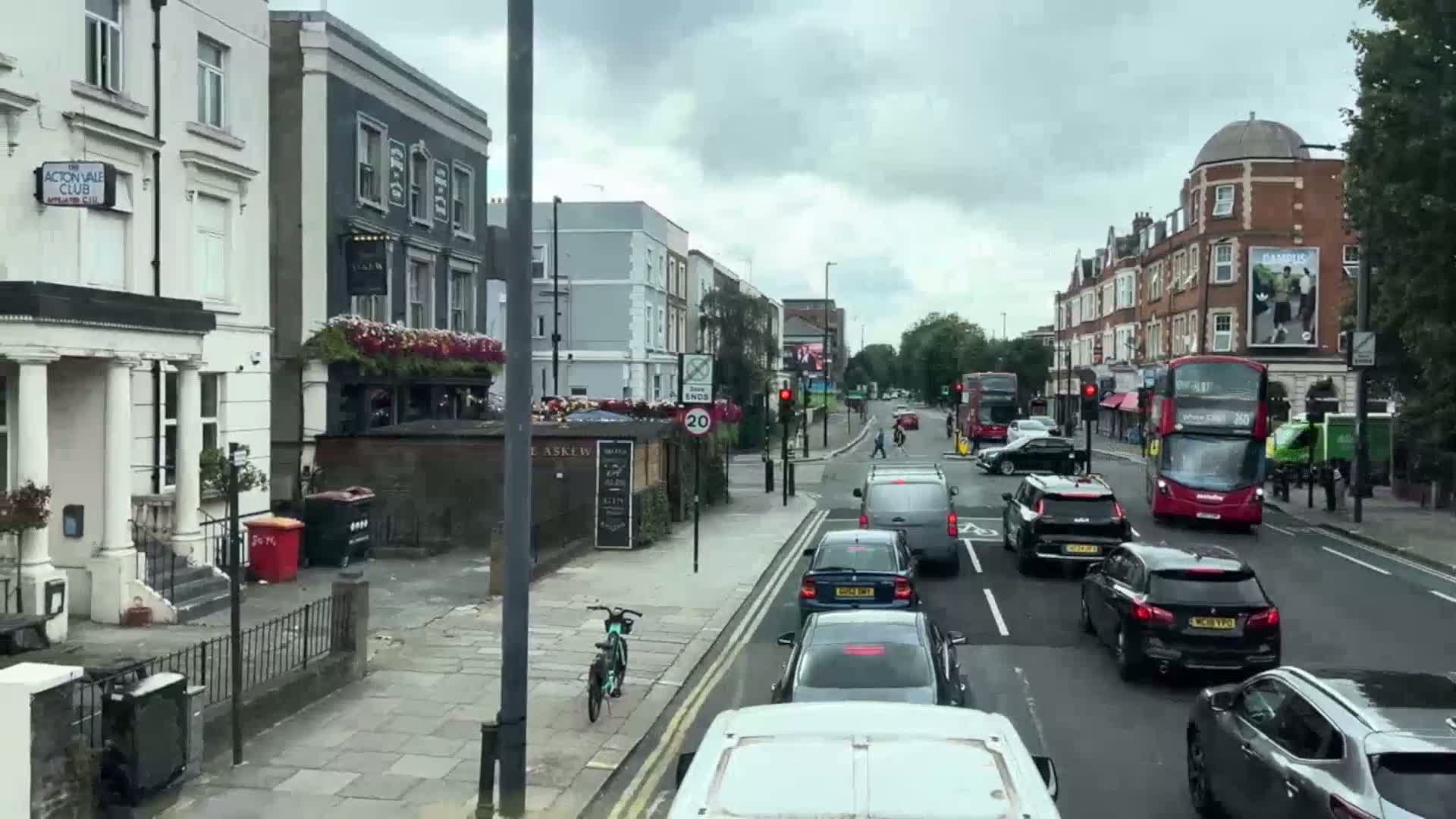 A cloudy day on a city street shows several cars stopped at an intersection, with double-decker buses visible in the distance and a bicycle parked on the sidewalk.