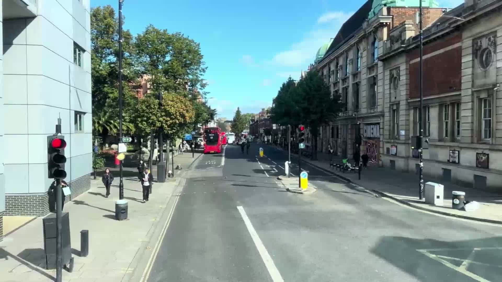 A sunny city street scene shows a red double-decker bus stopped at a traffic light, pedestrians walking on the sidewalk, and several buildings lining the street.