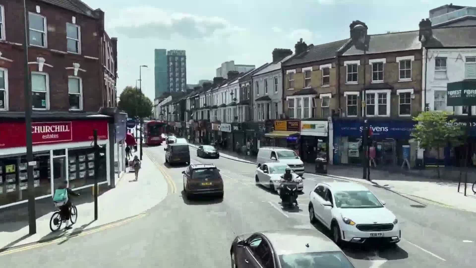 A sunny day on a city street with multiple cars, a bus, a motorcycle, and a cyclist traveling past shops and buildings.