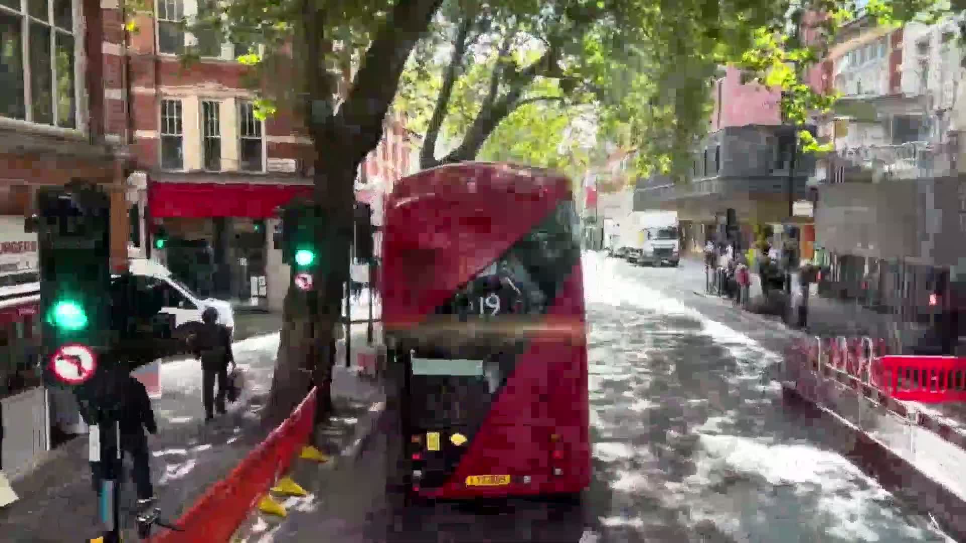 A red double-decker bus is stopped on a wet city street, surrounded by pedestrians and buildings, under a partly sunny sky.