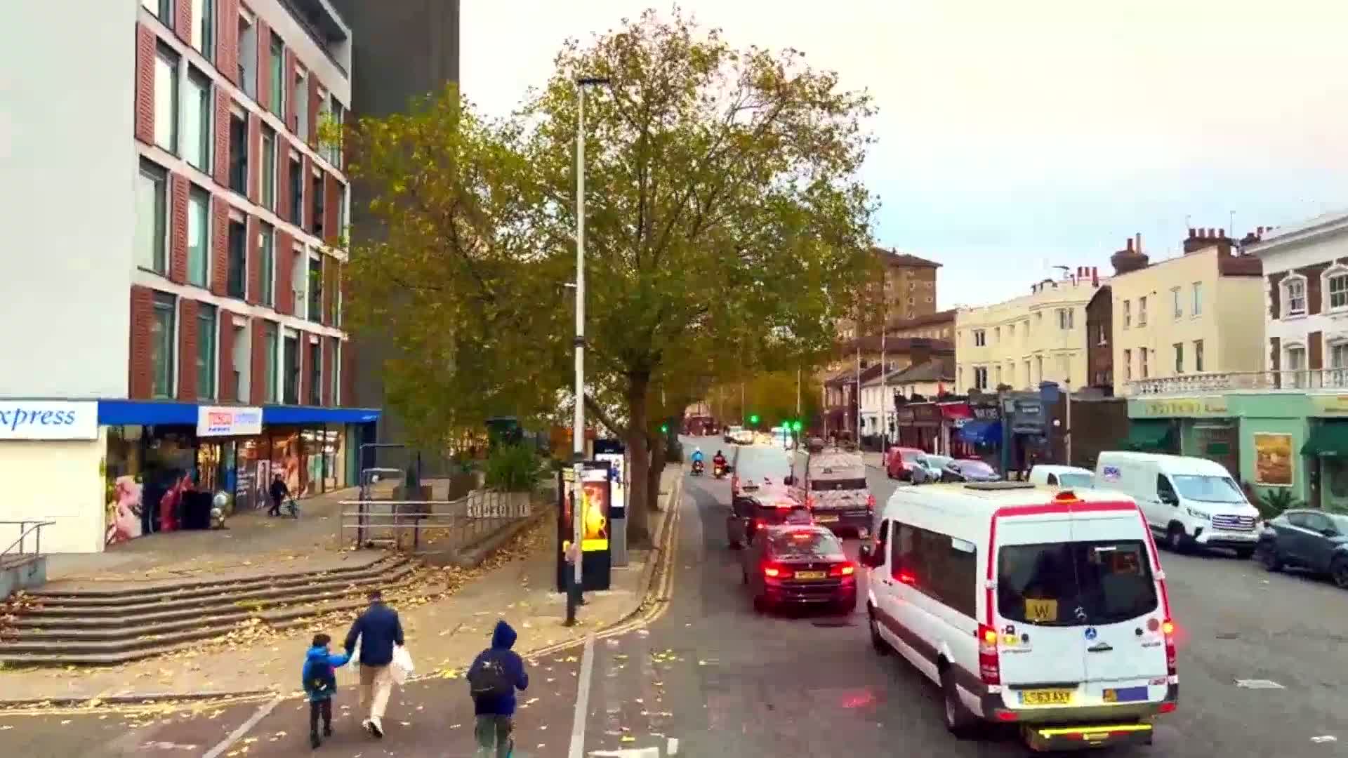 A city street scene on an overcast day shows a mix of modern and older buildings, with pedestrians and several vehicles including a minibus in traffic.