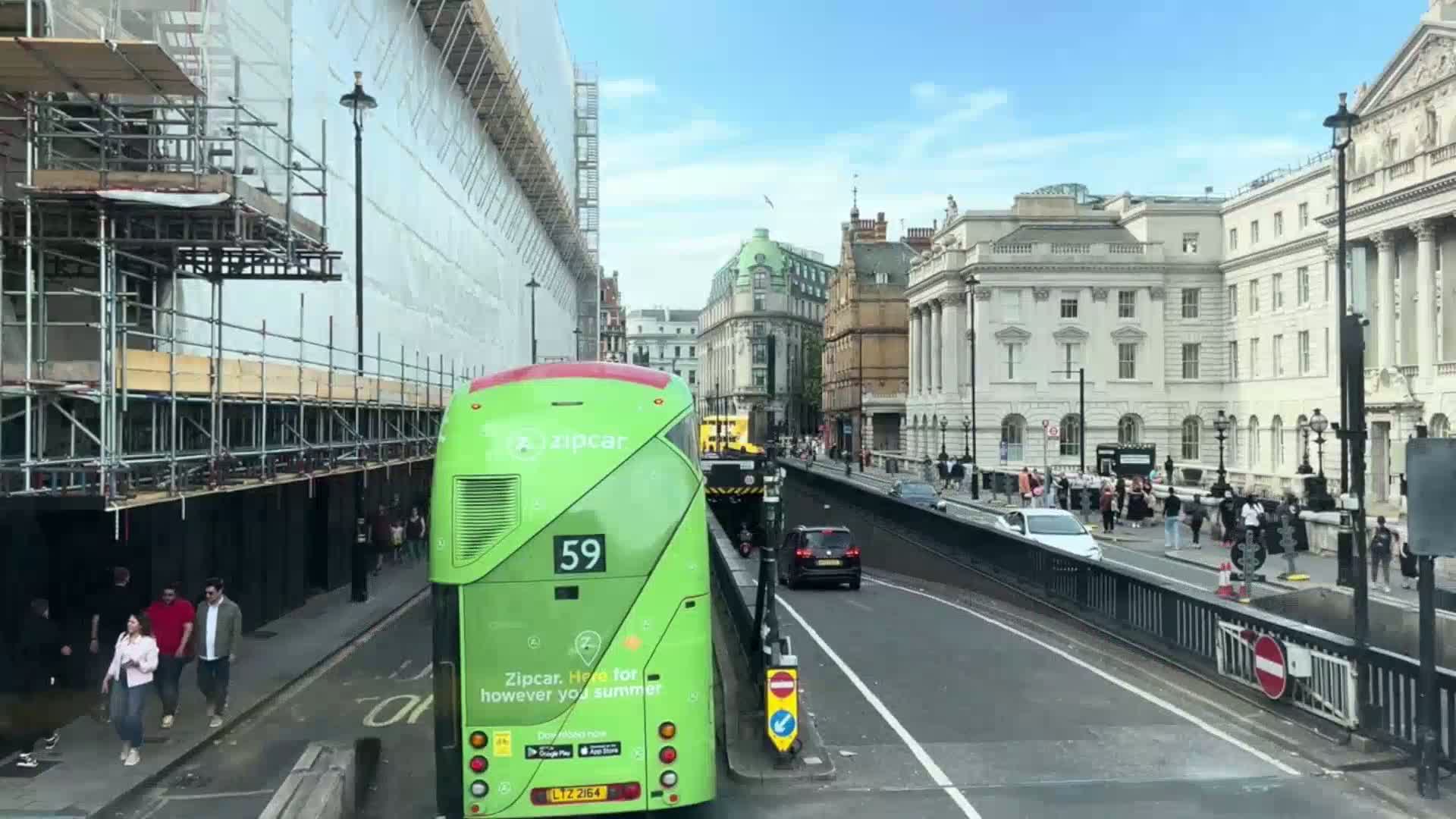 A lime green double-decker bus is stopped on a city street under a mostly sunny sky, with construction scaffolding visible on a building to the left and several pedestrians walking along the street and sidewalk.